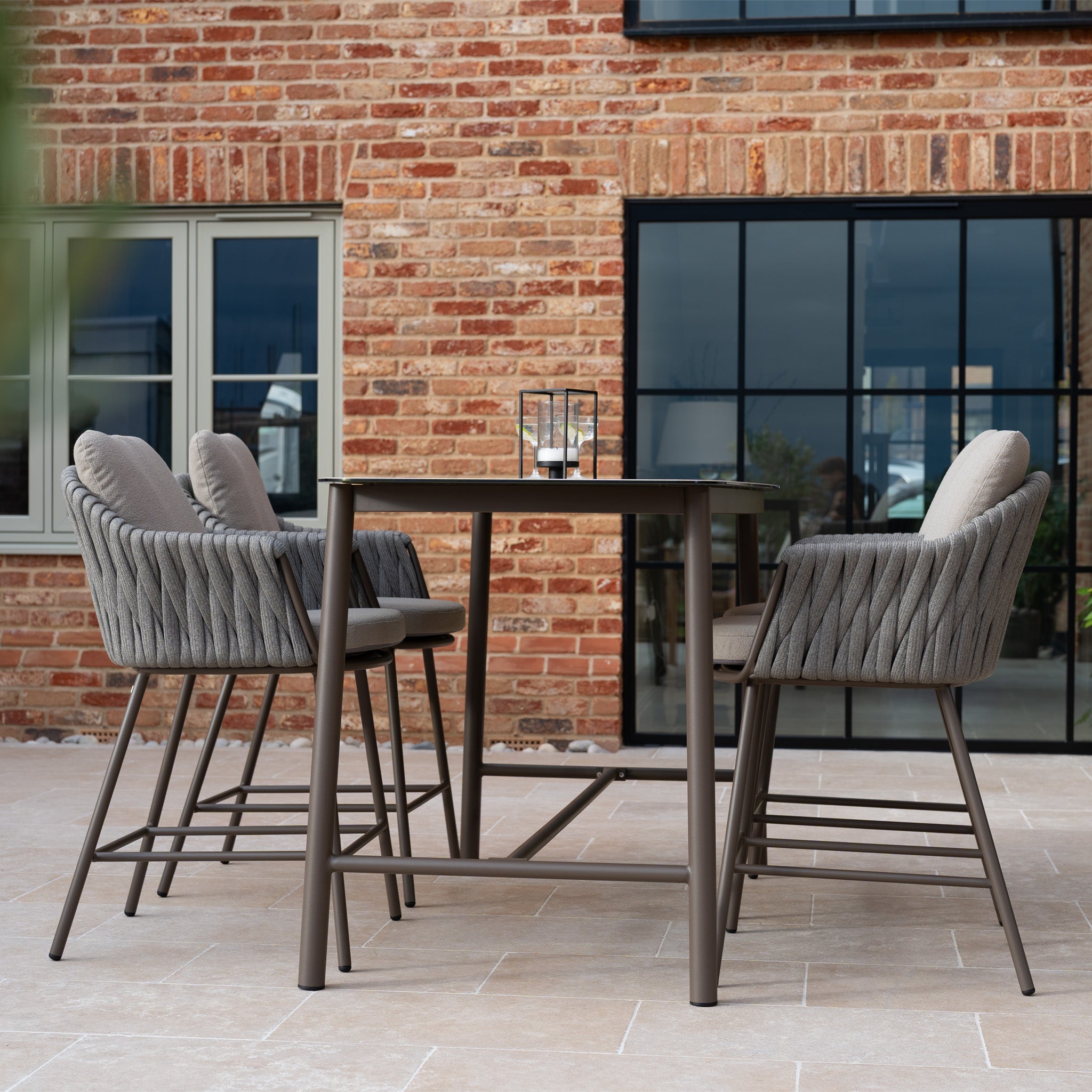 The Capri 4 Seat High Rectangular Dining Set in Stone Grey features stylish rope chairs and a metal table, ideal for patios with a brick wall.