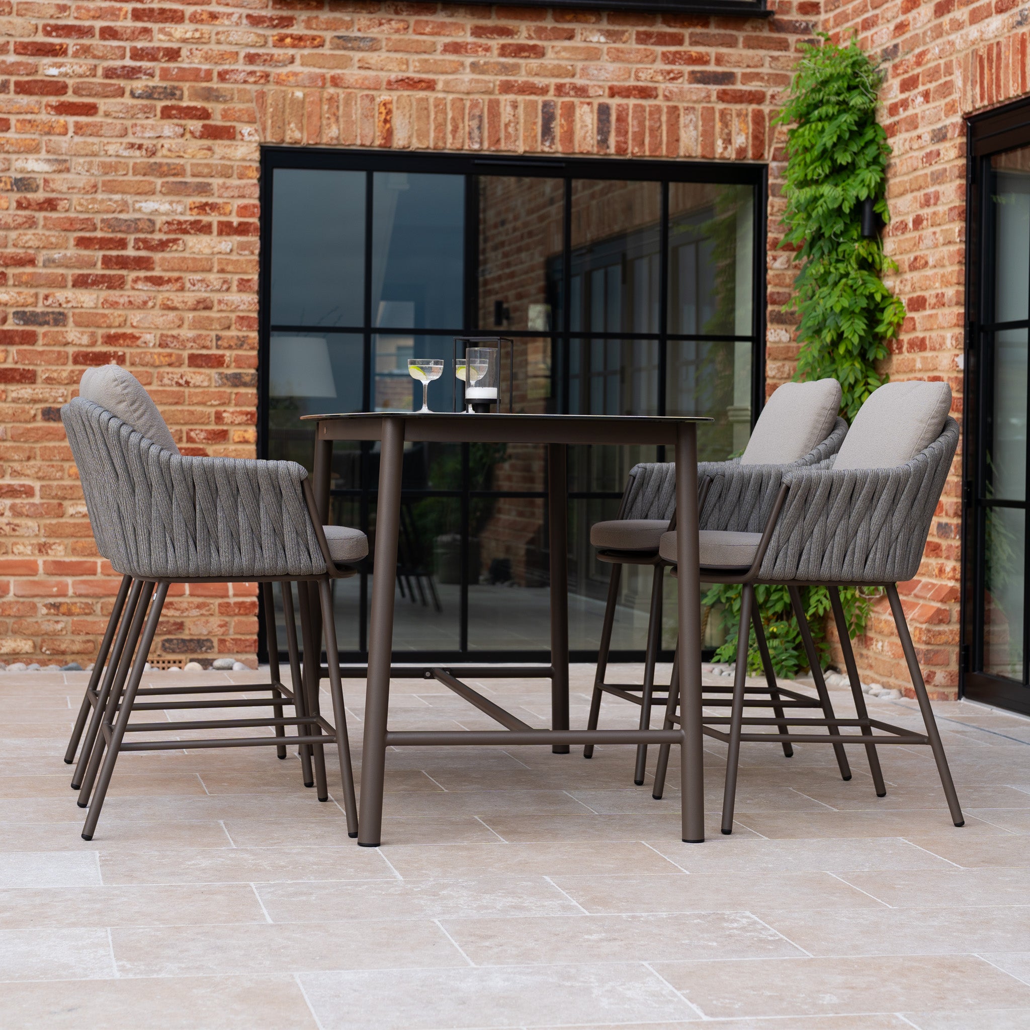 Outdoor patio with a Capri 4 Seat High Rectangular Dining Set in Stone Grey, cushioned chairs, and two glasses of water, set before a brick building for stylish outdoor dining.