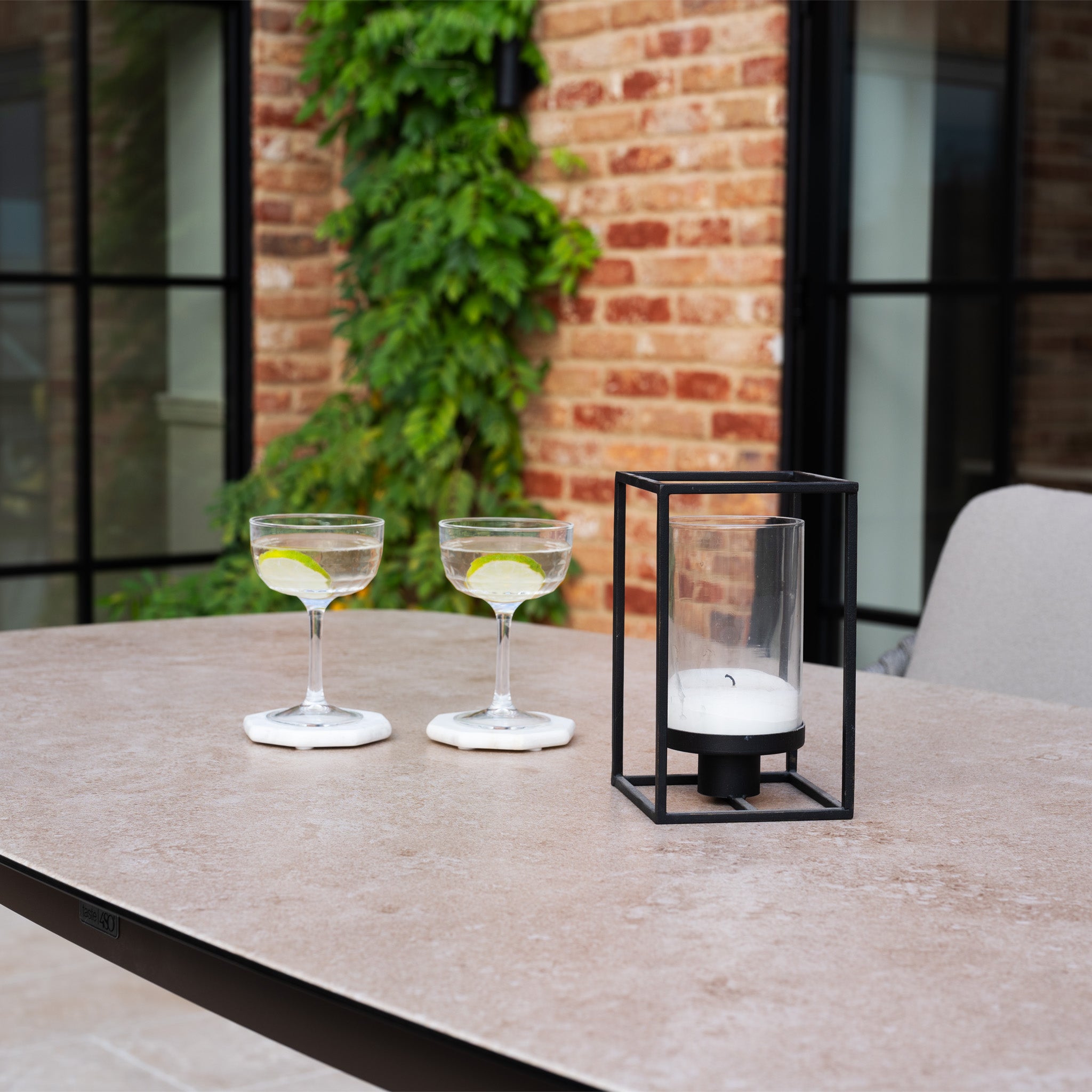Two lime cocktails and a candle lantern rest on an outdoor table, beautifully paired with the Capri 4 Seat High Rectangular Dining Set in Stone Grey near a brick wall and windows.