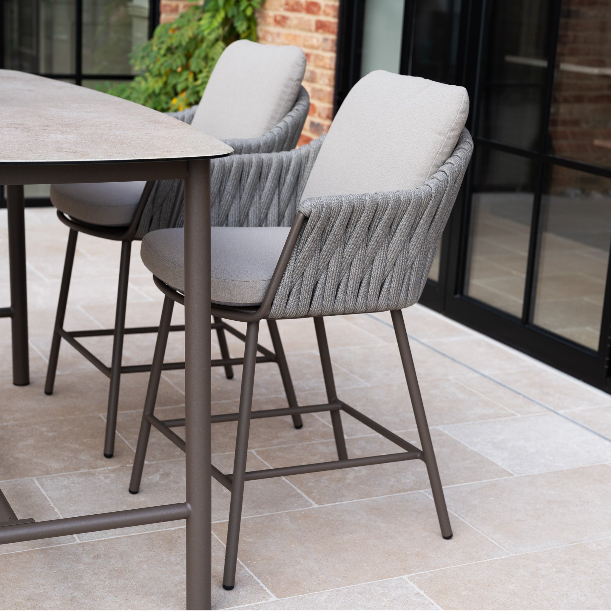 Two cushioned outdoor chairs with woven backs sit beside a table on a tiled patio near glass doors, creating a cozy spot that pairs perfectly with the Capri 4 Seat High Rectangular Dining Set in Stone Grey.