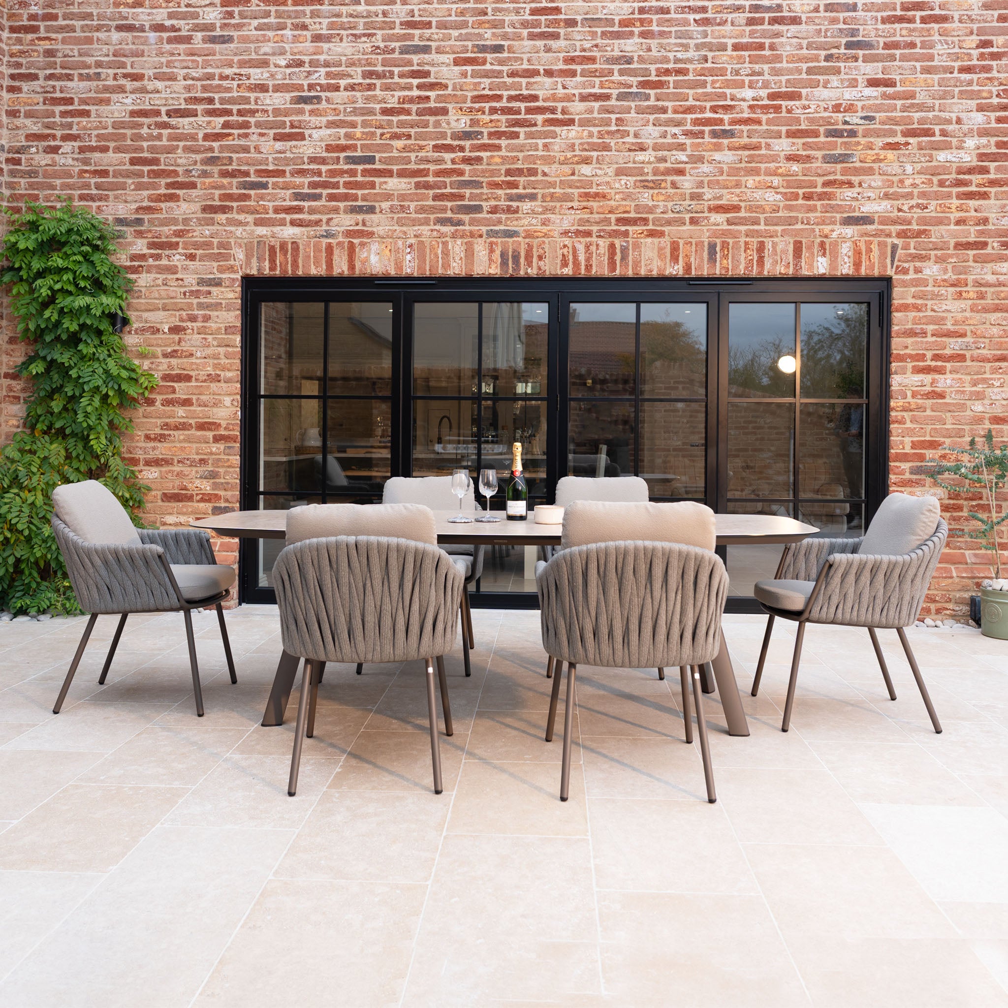 The Capri 6 Seat Curved Rectangular Dining Set in Stone Grey features hand-woven rope detailing, with six chairs arranged around a rectangular table—ideal for patios or outdoor spaces.