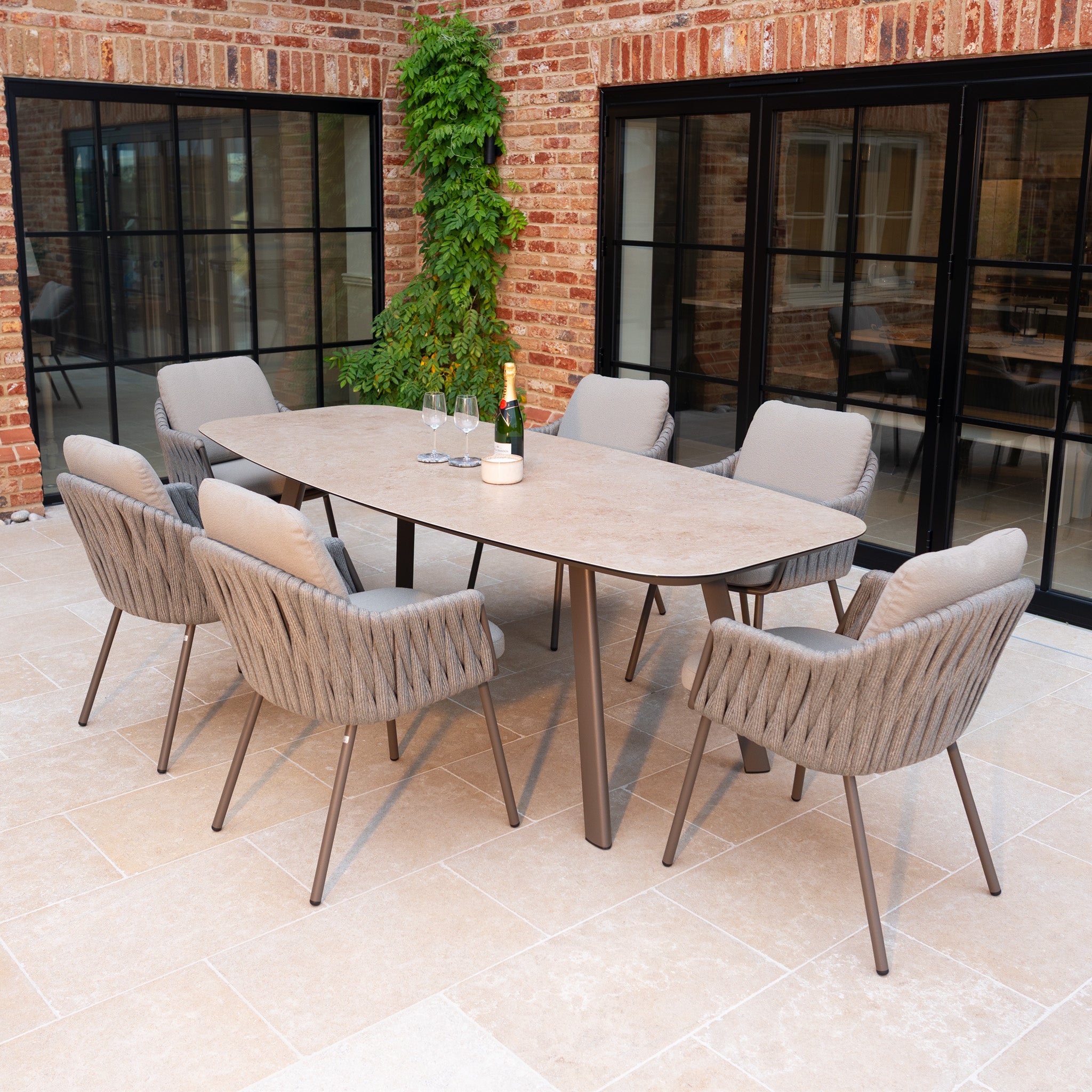 The Capri 6 Seat Curved Rectangular Dining Set in Stone Grey showcases hand-woven rope details, six cushioned chairs, and a sleek table—ideal for patios beside large windows and a brick wall.