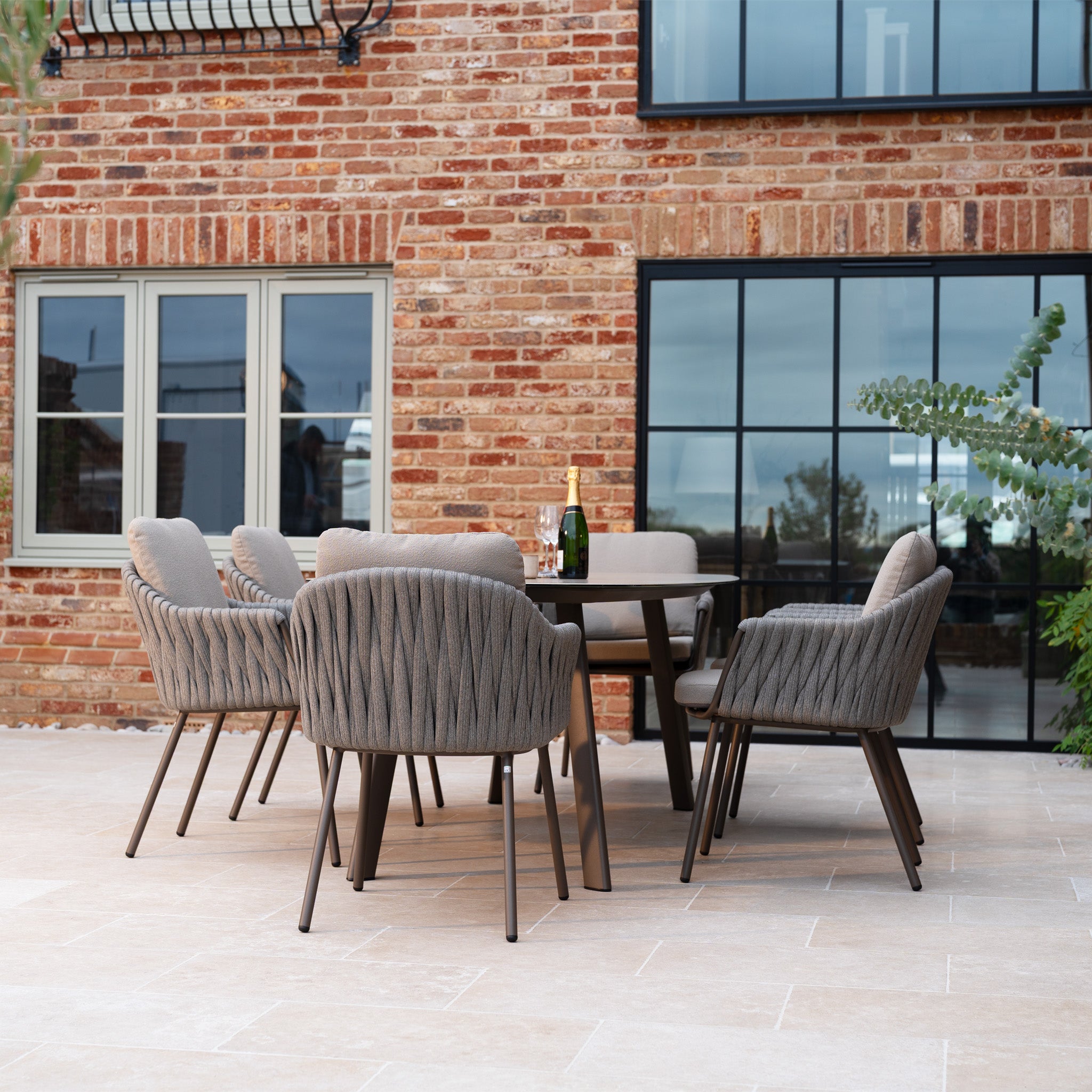 The Capri 6 Seat Curved Rectangular Dining Set in Stone Grey, with gray cushions and hand-woven rope accents, is arranged on a patio beside a brick building for stylish modern outdoor dining.