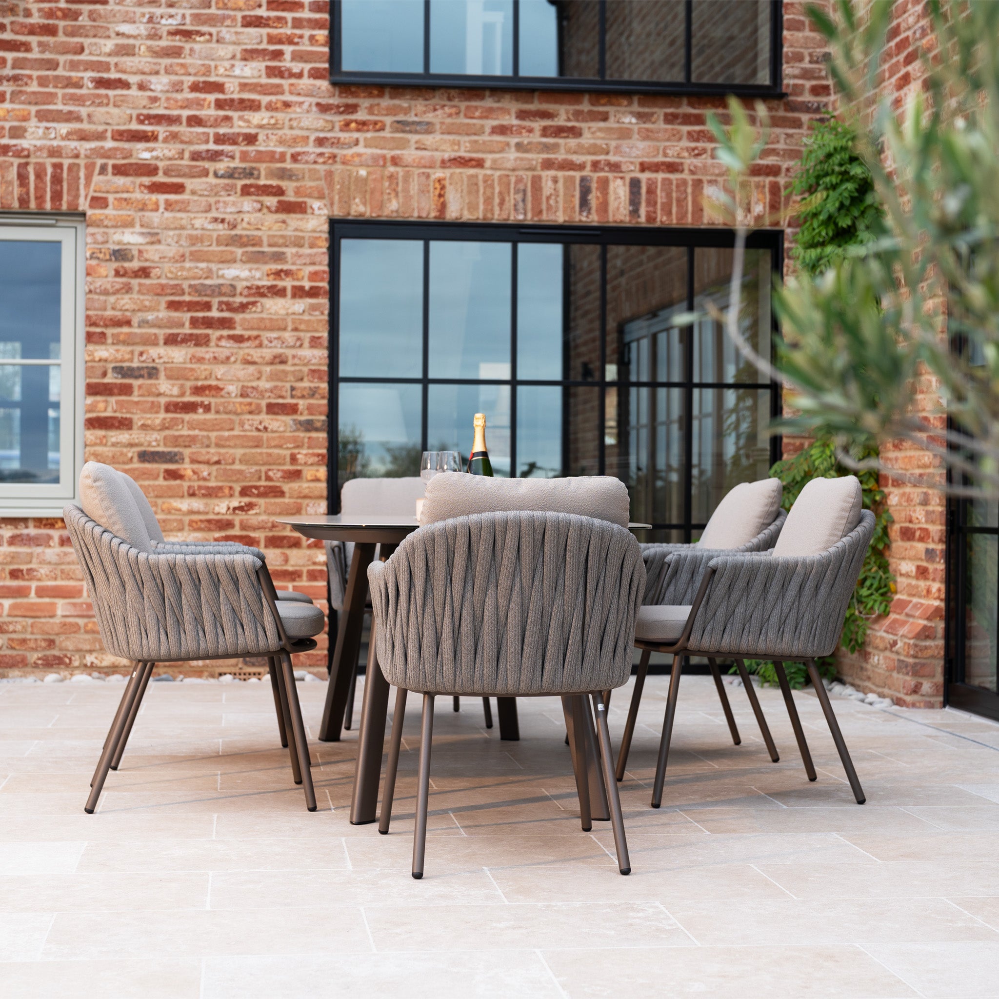 The outdoor patio showcases the Capri 6 Seat Curved Rectangular Dining Set in Stone Grey with cushioned chairs, hand-woven rope detailing, and a table, set with champagne and glasses against a brick building.