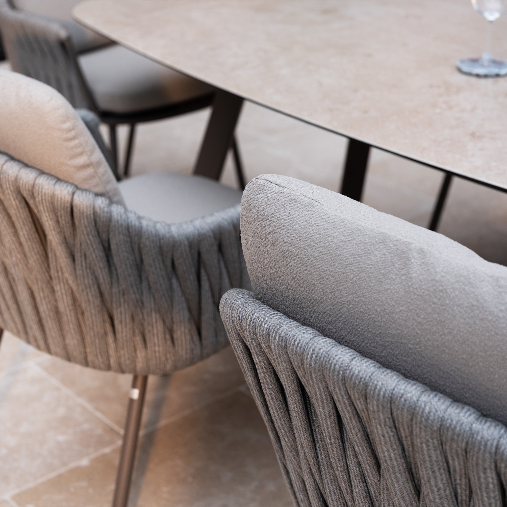 Close-up of cushioned gray chairs with hand-woven rope detailing surrounding a beige stone table on a tiled floor, featuring the elegant Capri 6 Seat Curved Rectangular Dining Set in Stone Grey.