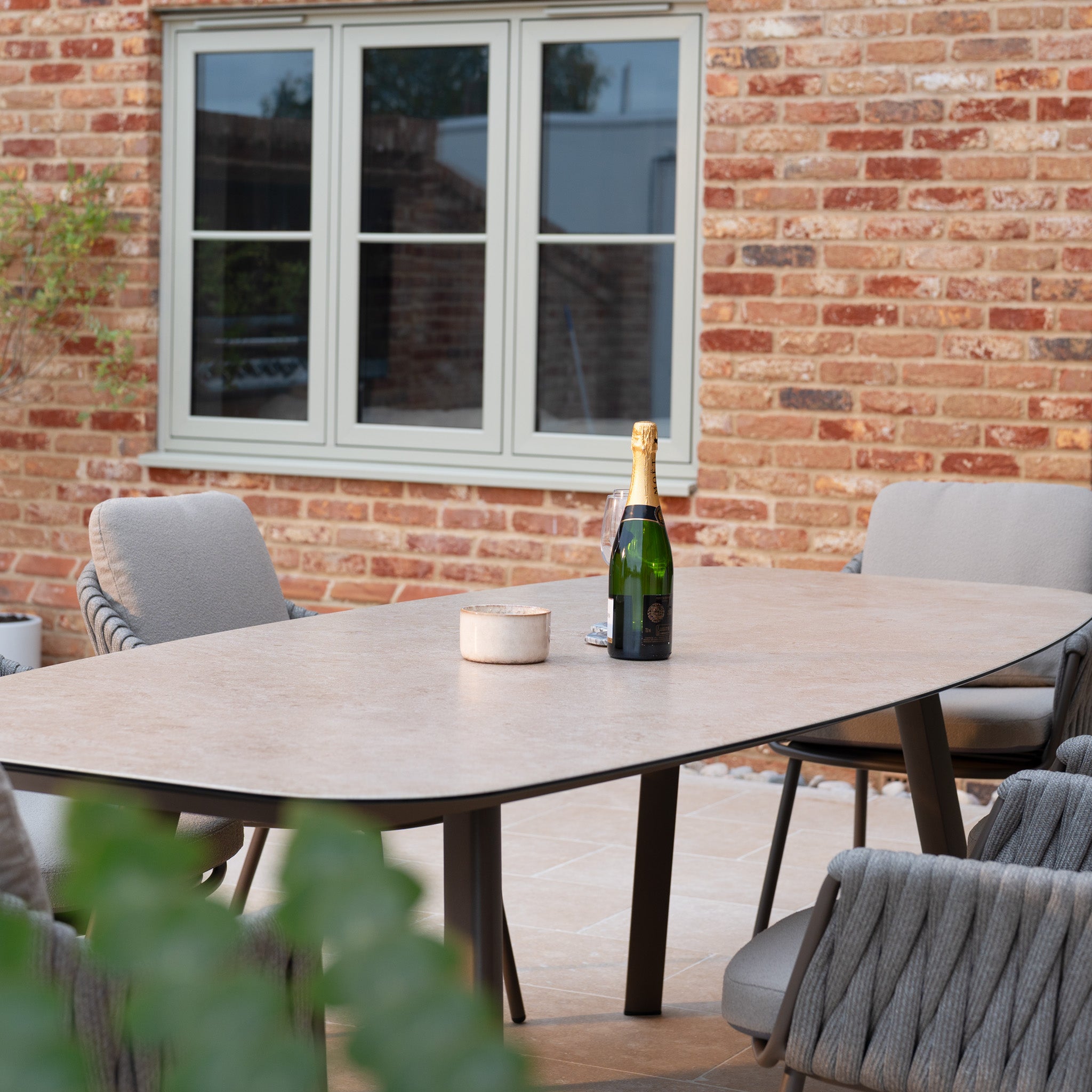 A champagne bottle and candle rest on the Capri 6 Seat Curved Rectangular Dining Set in Stone Grey, with chairs featuring hand-woven rope details, set outdoors near a brick wall and window.
