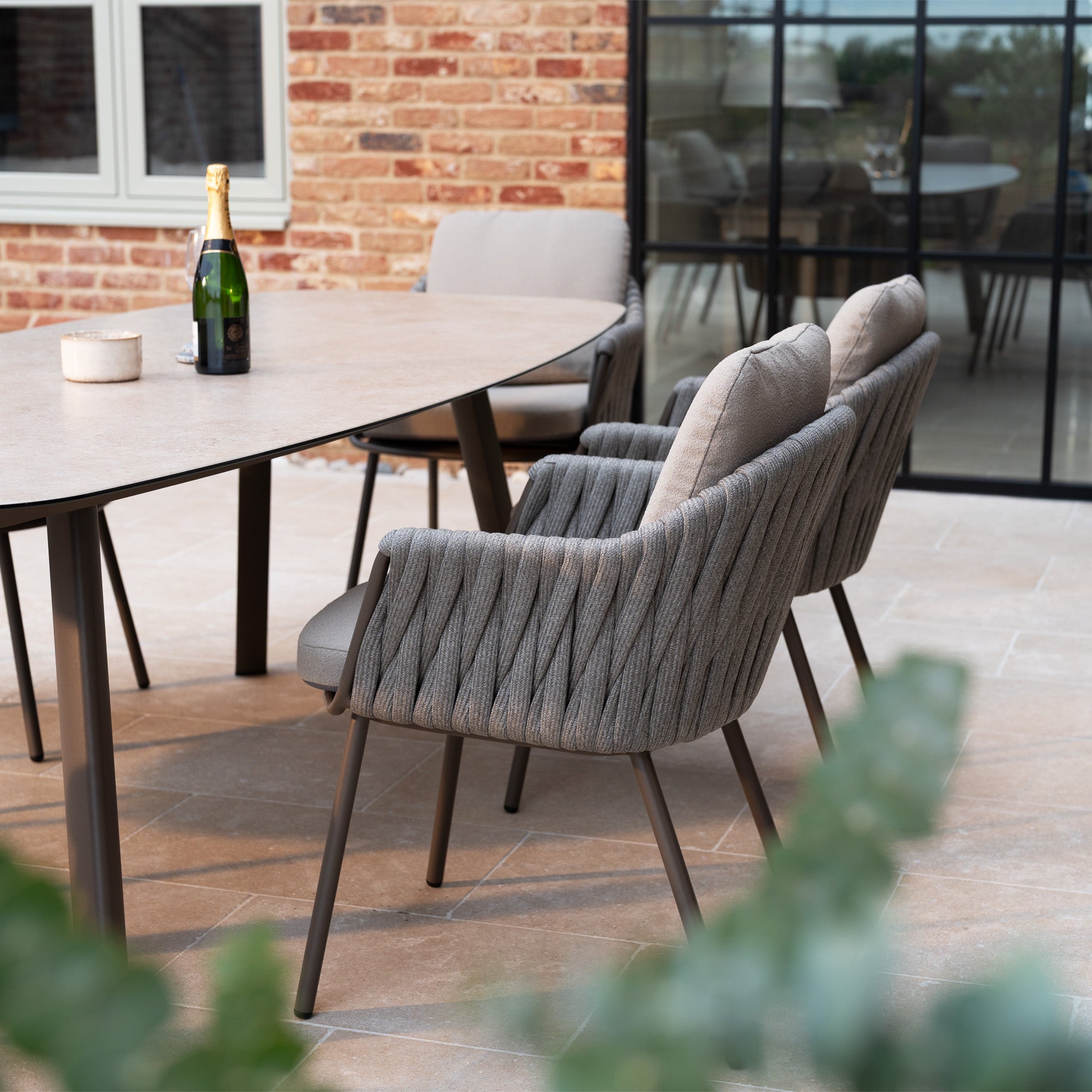Modern outdoor dining area featuring the Capri 6 Seat Curved Rectangular Dining Set in Stone Grey, with cushioned chairs, hand-woven rope accents, and a bottle of champagne on the table.