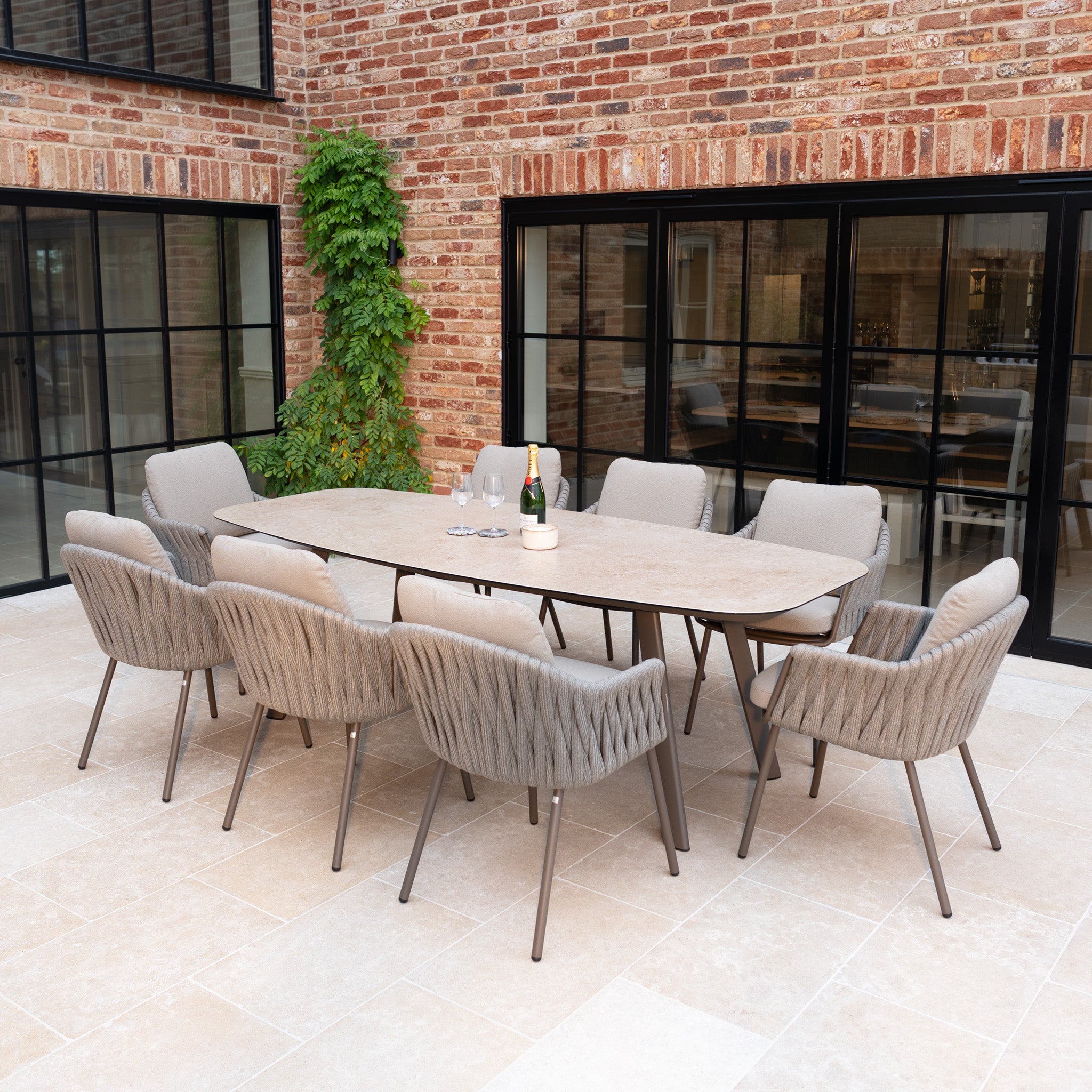 Host elegant outdoor meals beside a brick wall with the Capri 8 Seat Curved Rectangular Dining Set in Stone Grey, complete with champagne and glasses on a stone patio for a stylish garden experience.