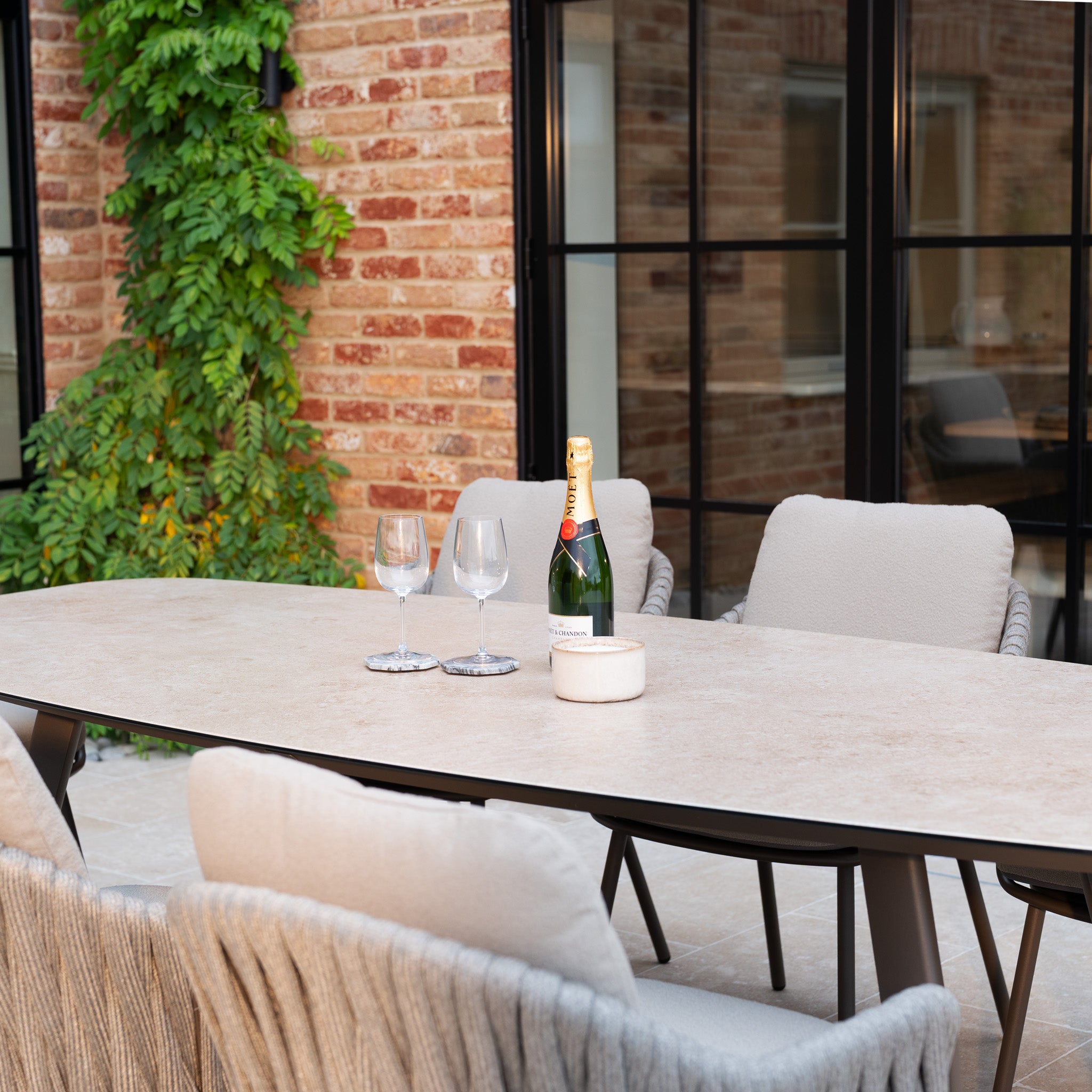 The Capri 8 Seat Curved Rectangular Dining Set in Stone Grey, shown with four chairs, a wine bottle, two glasses, and a candle by a brick wall and greenery, perfectly complements any outdoor space.