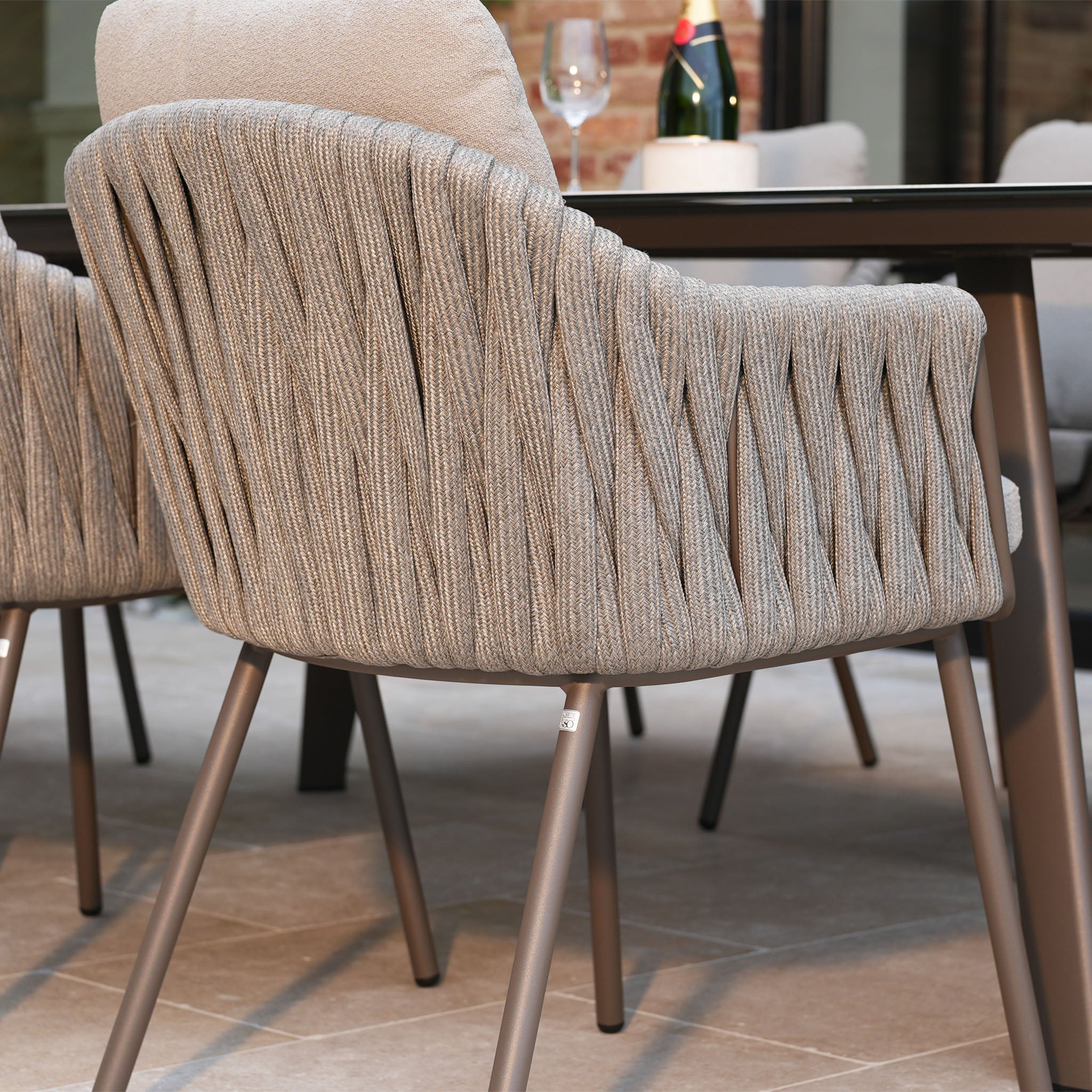 Close-up of a modern woven outdoor chair with metal legs beside a table on a tiled patio, part of the Capri 8 Seat Curved Rectangular Dining Set in Stone Grey.