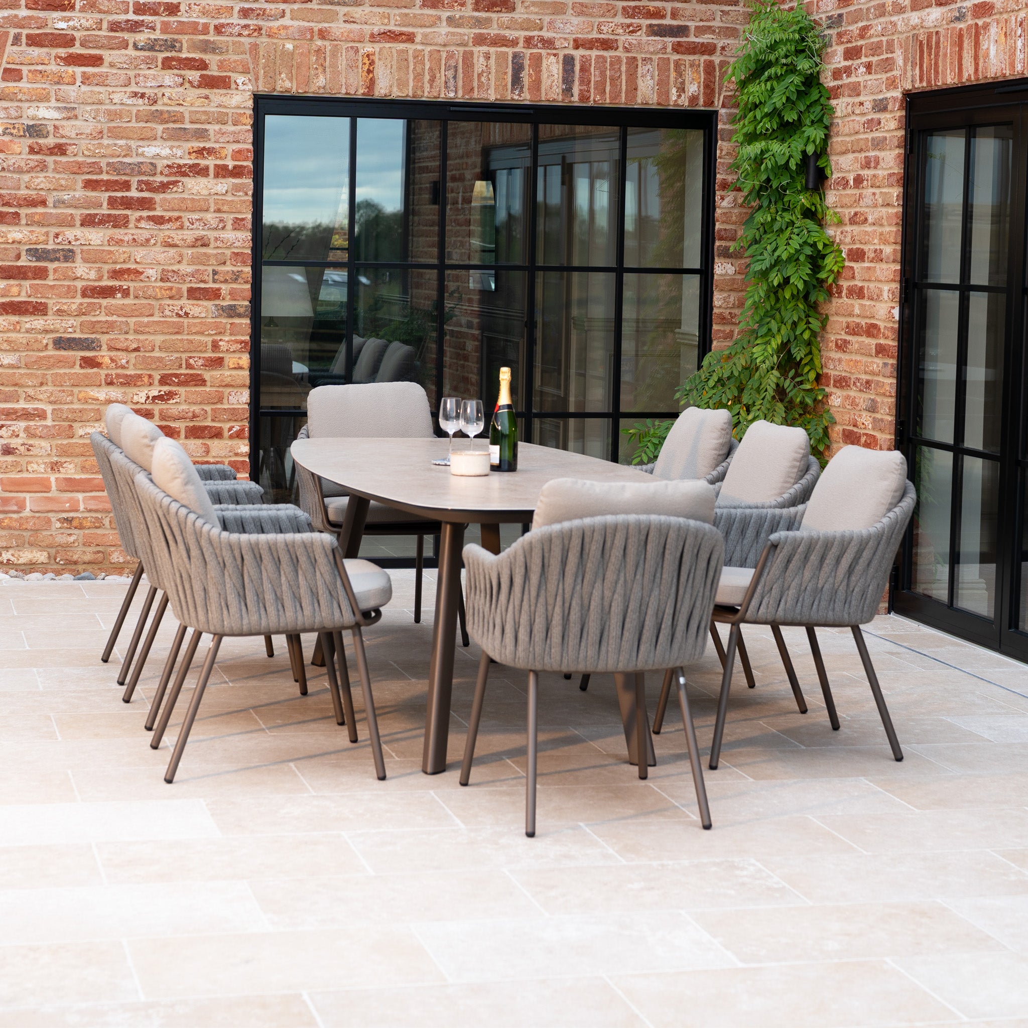 The Capri 8 Seat Curved Rectangular Dining Set in Stone Grey features cushioned chairs and a table, perfect for stylish outdoor gatherings on a patio with wine glasses and a bottle by brick walls.