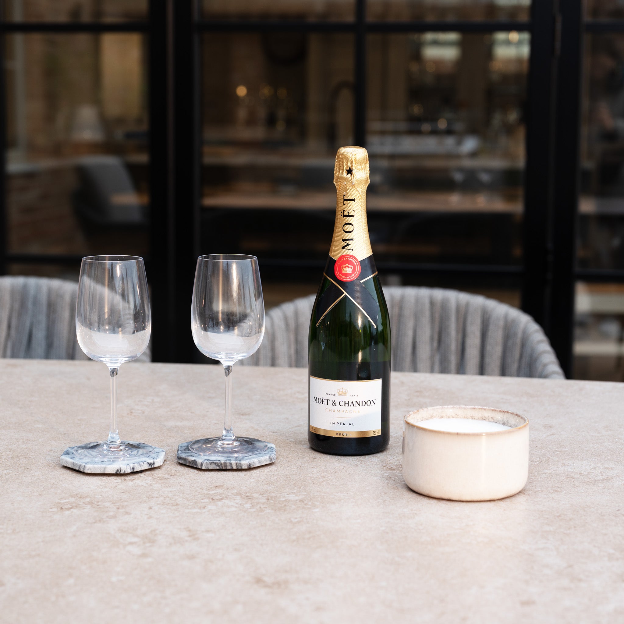 A bottle of Moët & Chandon, two empty wine glasses, and a ceramic bowl sit atop the Capri 8 Seat Curved Rectangular Dining Set in Stone Grey.