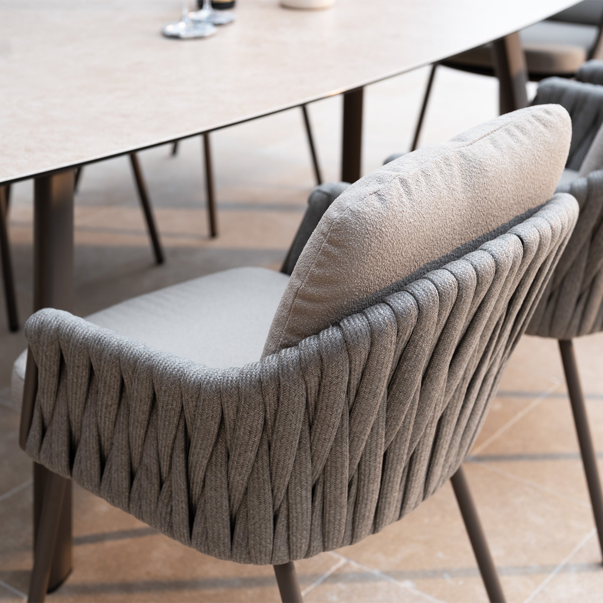 Close-up of a modern cushioned chair with woven sides from the Capri 8 Seat Curved Rectangular Dining Set in Stone Grey, placed beside a light table on tiled flooring—ideal elegant garden furniture for outdoor dining.