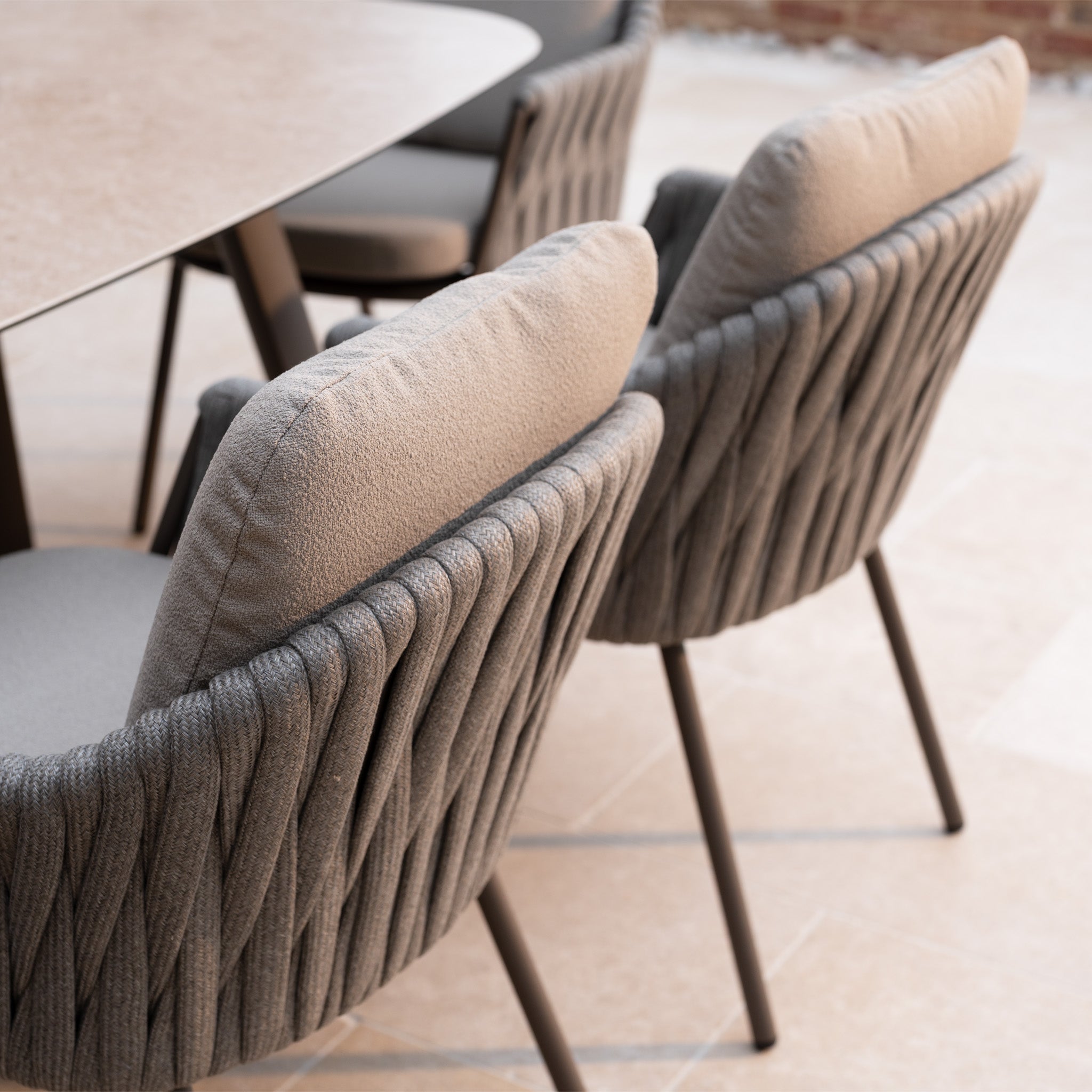 Two cushioned, grey woven chairs from the Capri 8 Seat Curved Rectangular Dining Set in Stone Grey are placed by a table on a light stone floor, creating an inviting and stylish outdoor dining space.
