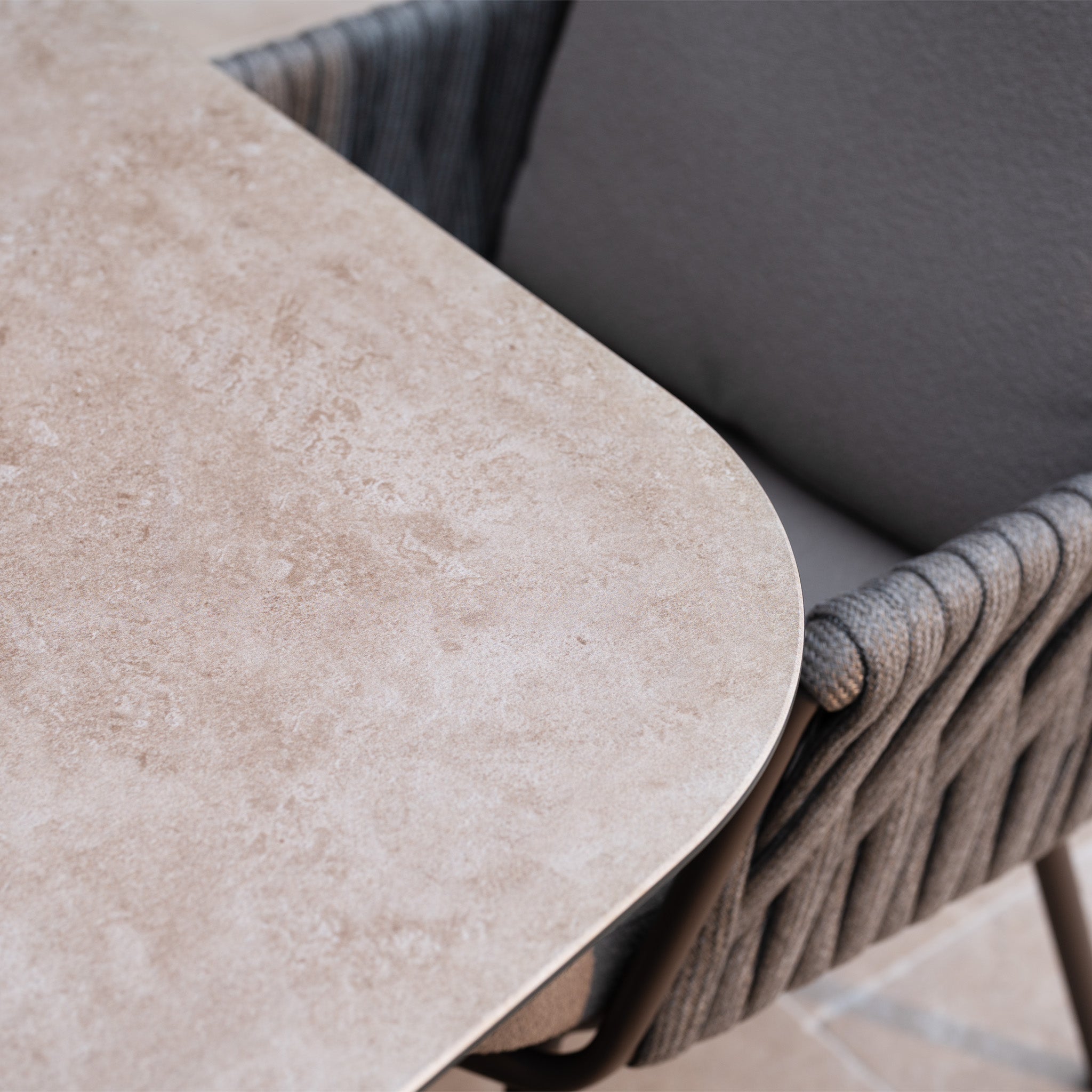 Close-up of a beige stone tabletop beside a gray cushioned chair from the Capri 4 Seat High Rectangular Dining Set in Stone Grey, highlighting its woven texture—ideal for stylish outdoor dining.