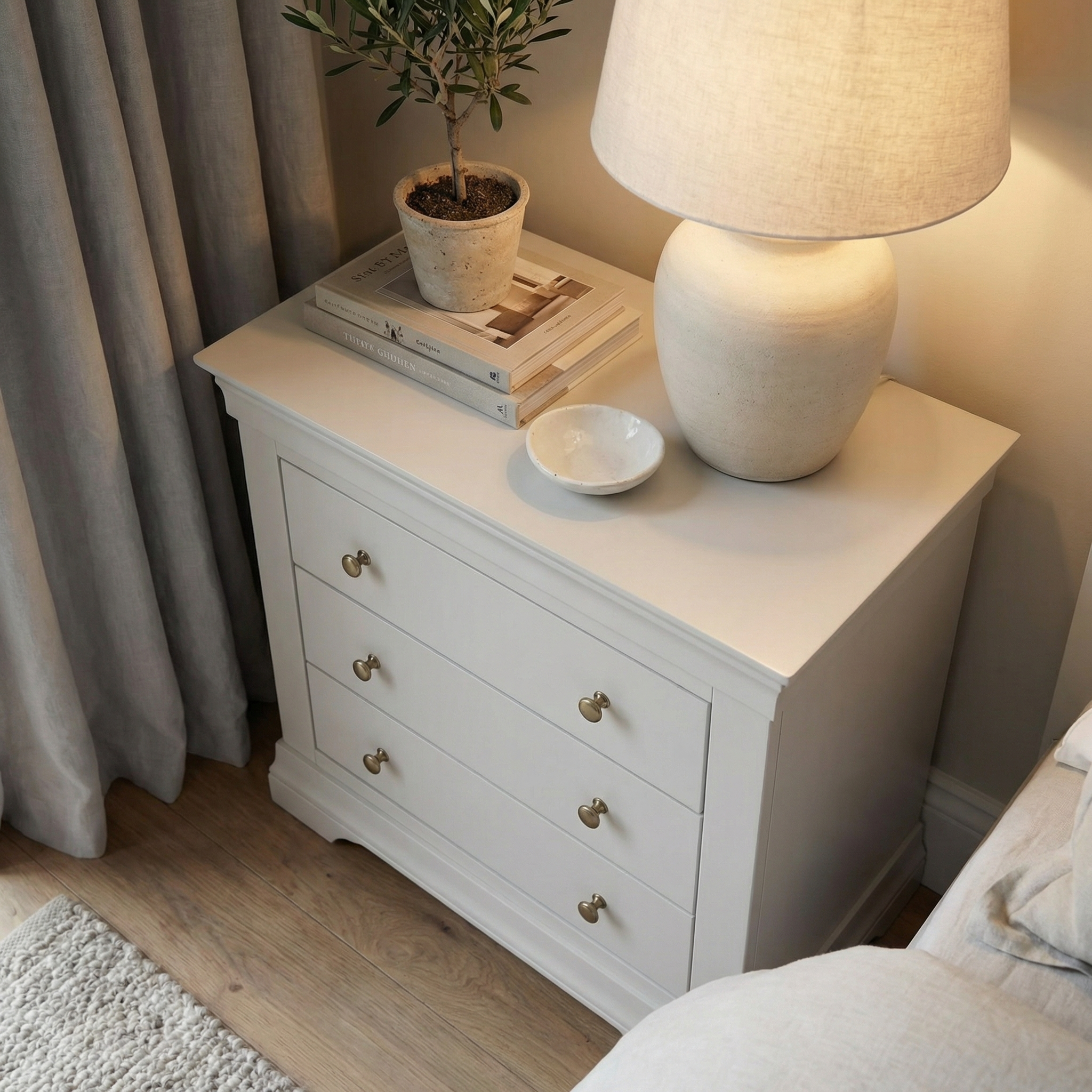 A white nightstand with books, a potted plant, a ceramic lamp, and a bowl sits beside the bed and curtains, complemented by the elegant Chalbury 3 Drawer Chest in Pebble Grey for added style and storage.