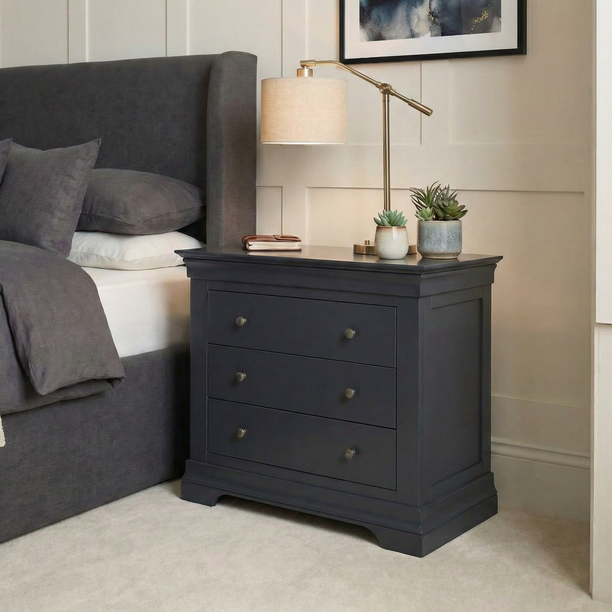 A Chalbury 3 Drawer Chest in Dusky Black serves as a modern storage piece, topped with a lamp, books, and small plants beside a bed dressed in gray bedding.