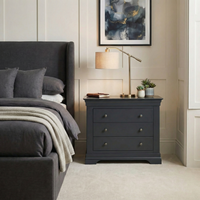 Modern bedroom featuring the Chalbury 3 Drawer Chest in Dusky Black as a nightstand, paired with a lamp, plants, and abstract art above, next to a gray upholstered bed—ideal modern storage for a sleek look.