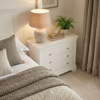 A cozy bedroom with a white nightstand, lamp, books, plant, and a bed with knitted blankets features the Chalbury 3 Drawer Chest in Warm White—contemporary storage that adds style and function.