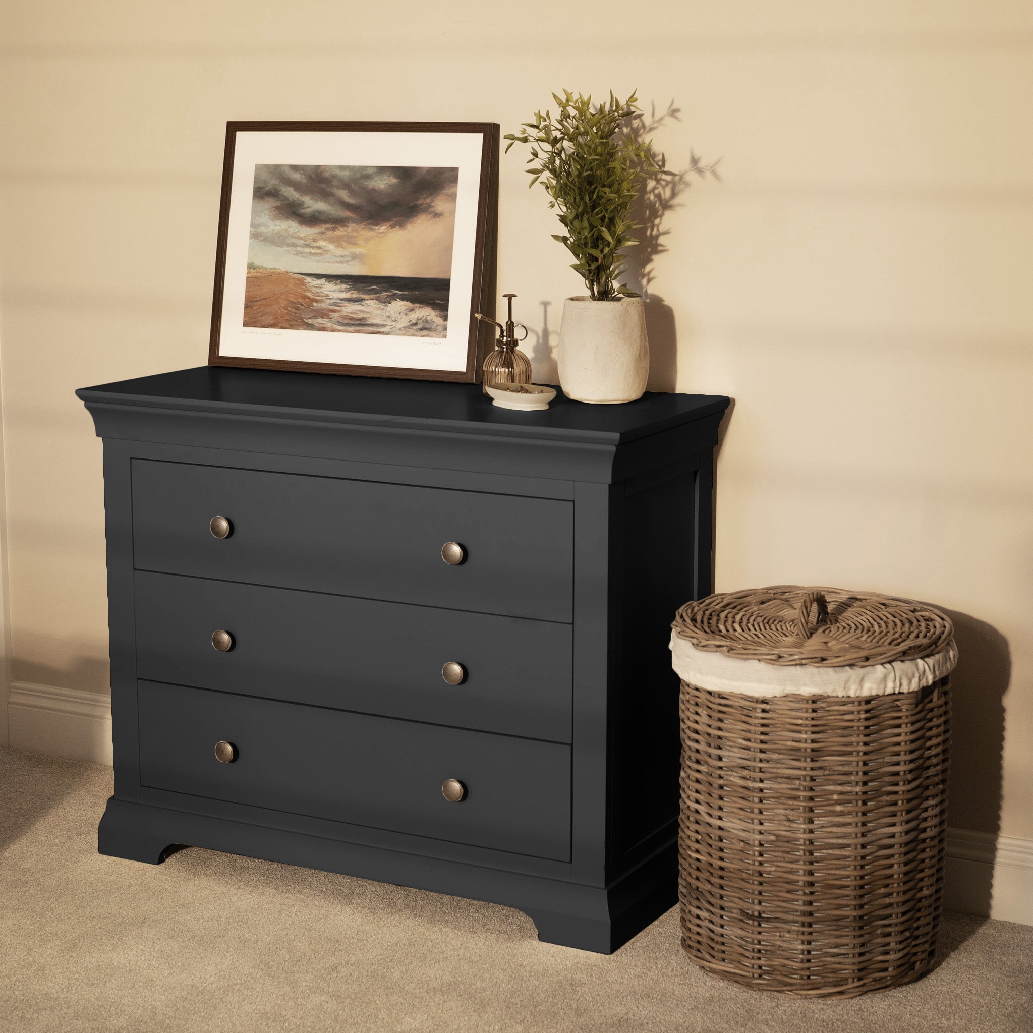 A Chalbury 3 Drawer Chest in Dusky Black offers modern storage, styled with a framed seascape, greenery, and a wicker laundry basket nearby.