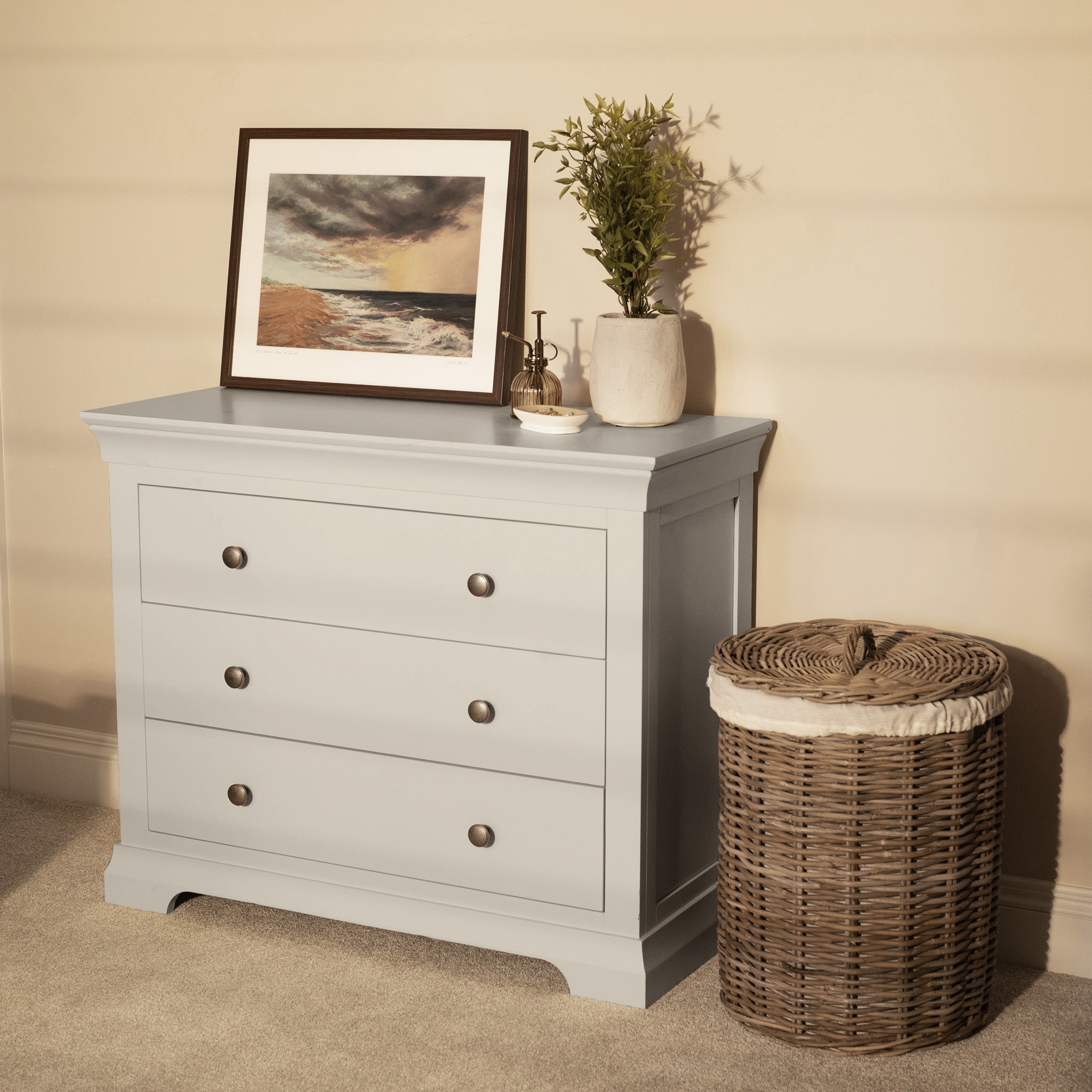 Pebble Grey Chalbury 3 Drawer Chest with three drawers, topped with a framed seascape, vase, and basket, sits on a beige carpeted floor.