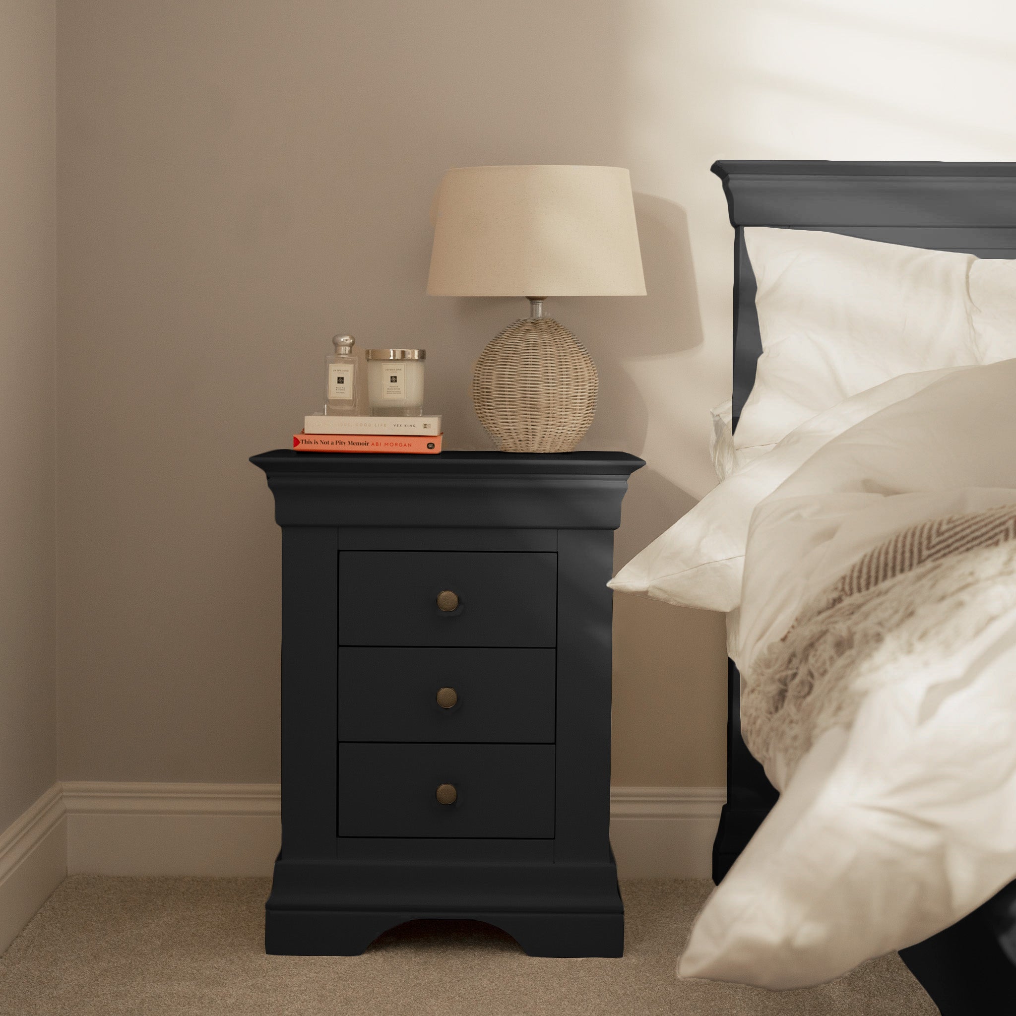 The Chalbury 3 Drawer Bedside Table in Dusky Black offers ample storage with three drawers, ideal for keeping a lamp, books, and perfume bottles organized beside a neatly made bed.