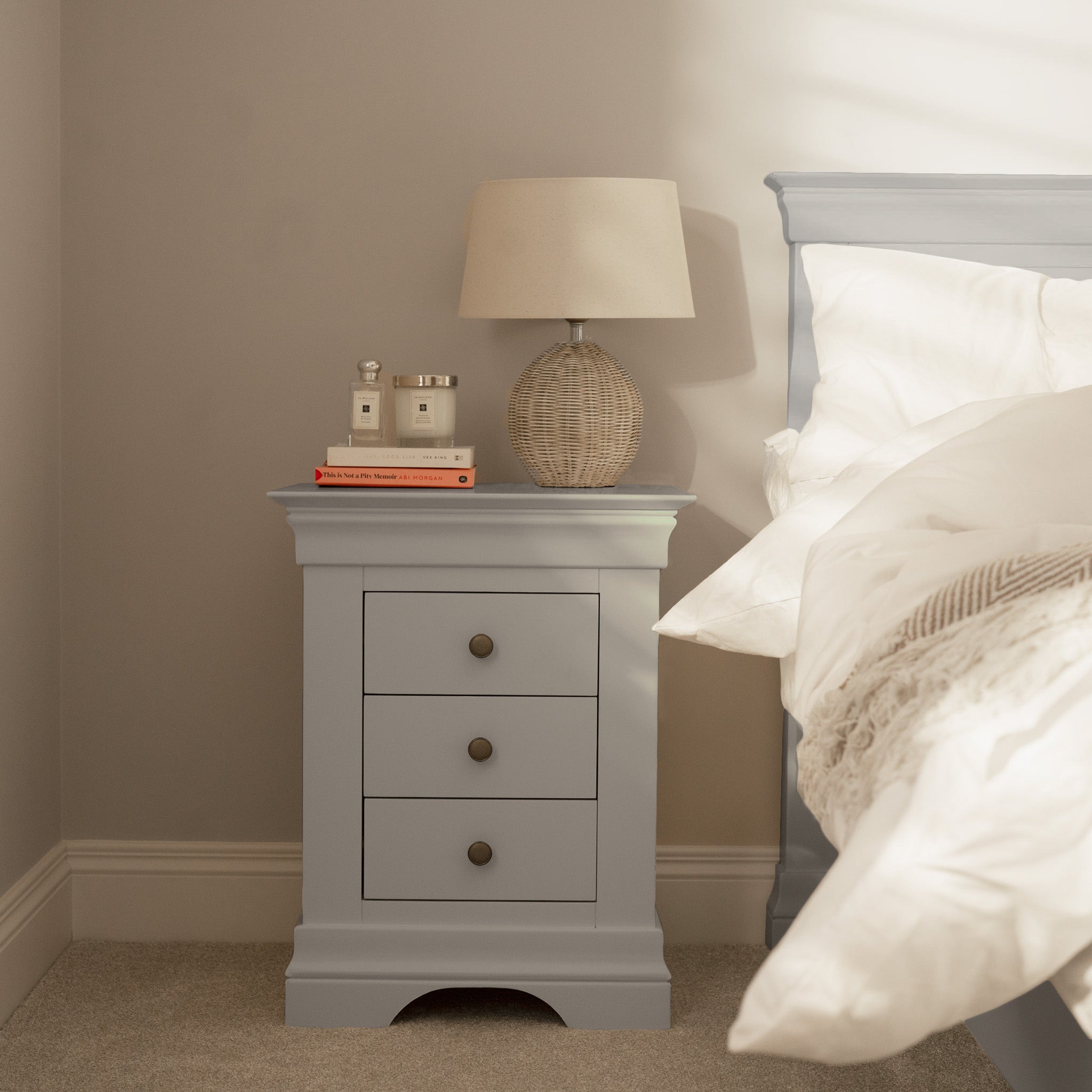 The Chalbury 3 Drawer Bedside Table in Pebble Grey placed next to a bed with white bedding, styled with a lamp, books, and decor, brings a modern touch to your room.