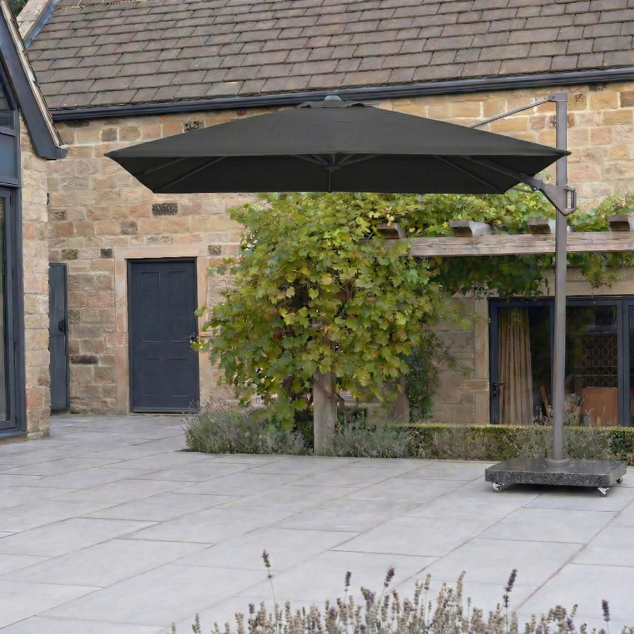 The Pacific Lifestyle Challenger T2 3m Square Cantilever Parasol in Anthracite shades a tiled courtyard beside a stone house with green plants.