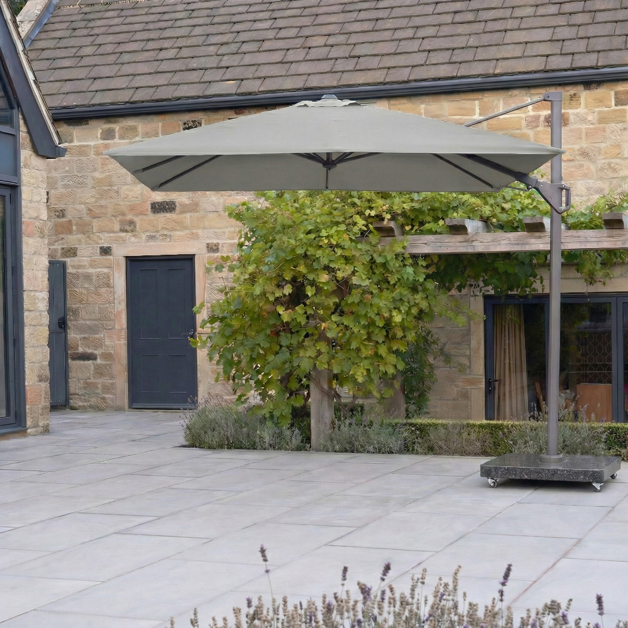 The Pacific Lifestyle Challenger T2 3m Square Cantilever Parasol in Grey stands on a tiled outdoor area near a stone building with green leafy vines.