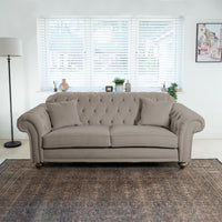 The Chesterfield 3 Seat Velvet Sofa in Mink, with its tufted details and cushions, elegantly enhances a bright living room. It is beautifully positioned near a window with blinds and complemented by a stylish floor lamp.