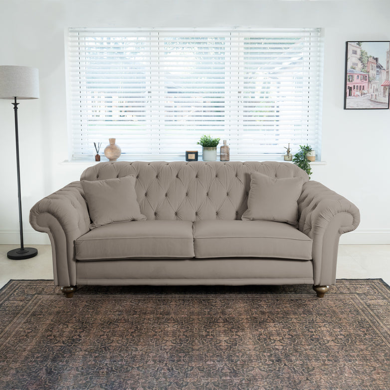 The Chesterfield 3 Seat Velvet Sofa in Mink, with its tufted details and cushions, elegantly enhances a bright living room. It is beautifully positioned near a window with blinds and complemented by a stylish floor lamp.