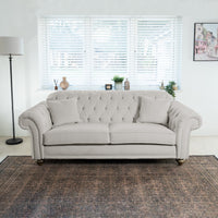 A Chesterfield 3 Seat Velvet Sofa in Feather Grey, adorned with cushions, sits in a bright living room, enhanced by a standing lamp and decor on the windowsill.