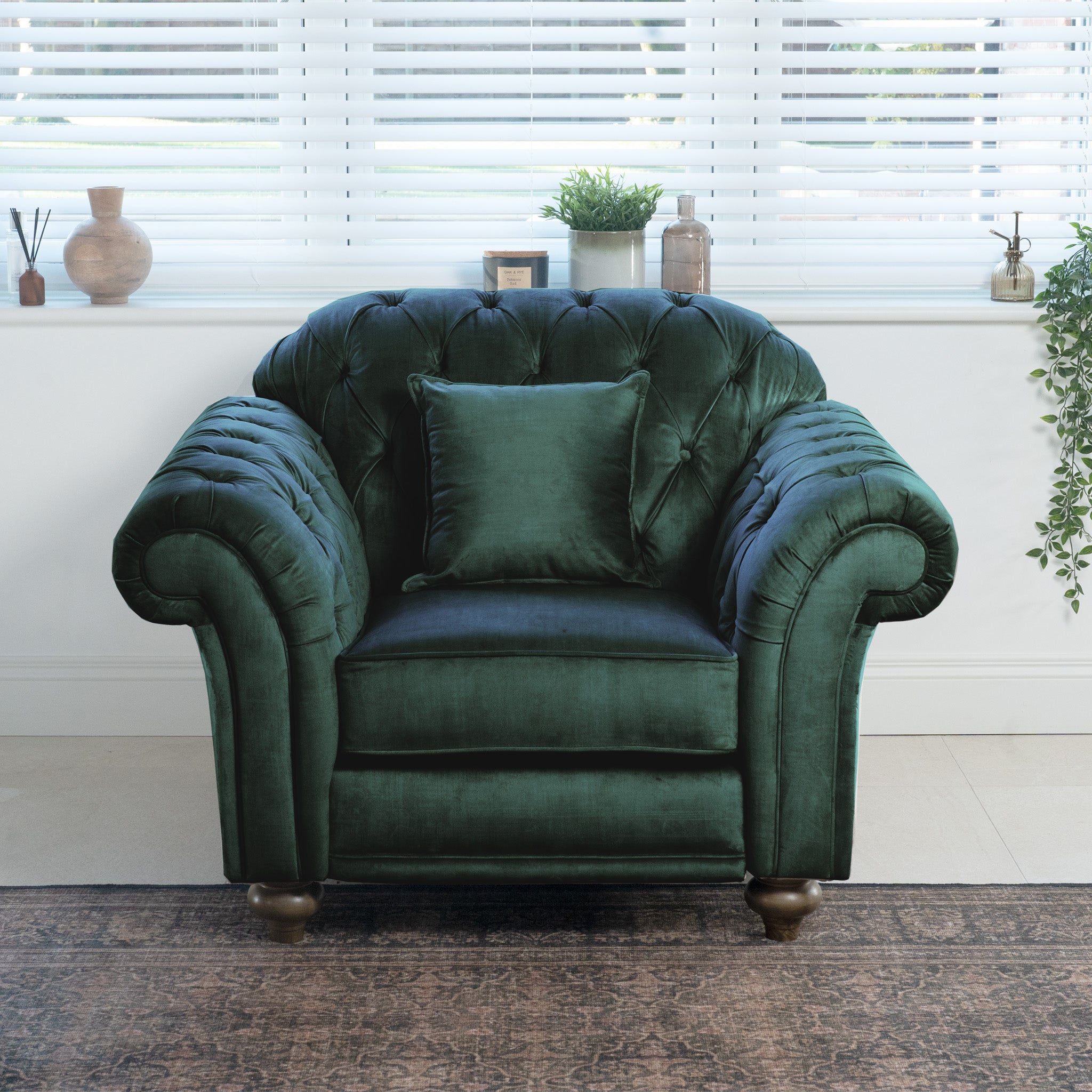 A Chesterfield Velvet Armchair in Bottle Green graces the bright room with its handcrafted elegance, accompanied by a matching cushion. The deep, alluring color is beautifully highlighted by white blinds and tasteful decorative elements, infusing the space with classic English charm.