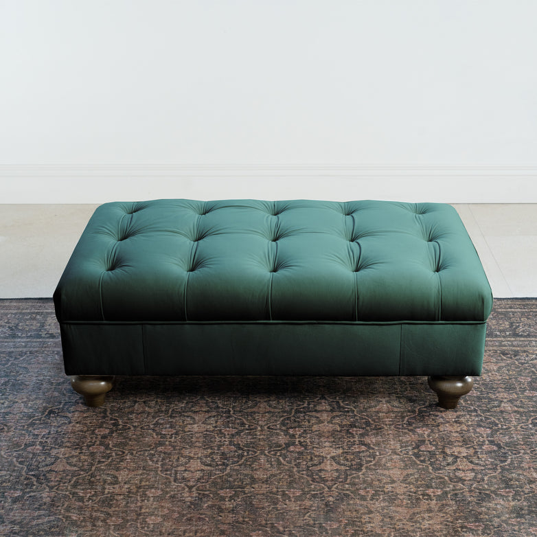 The Chesterfield Velvet Footstool in Bottle Green sits elegantly on a patterned rug in a minimalist room, adding a touch of sophistication.