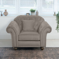 The Chesterfield Velvet Armchair in Mink, handcrafted in England with gray plush velvet upholstery, sits elegantly in front of a window. It's adorned with a cushion, surrounded by plants and vases, all placed on a patterned rug.