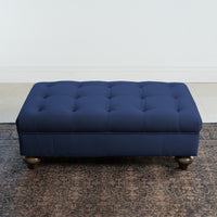 The Chesterfield Velvet Footstool in Royal Blue enhances a minimalist room with its luxurious presence atop a patterned rug.