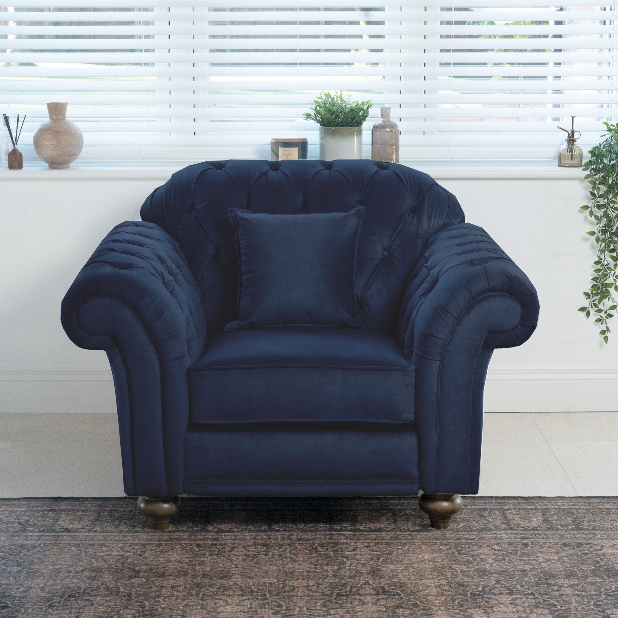 A Chesterfield Velvet Armchair in Royal Blue with a matching cushion adorns a bright room filled with plants and decor on the windowsill, elegantly blending comfort and style into the serene setting.