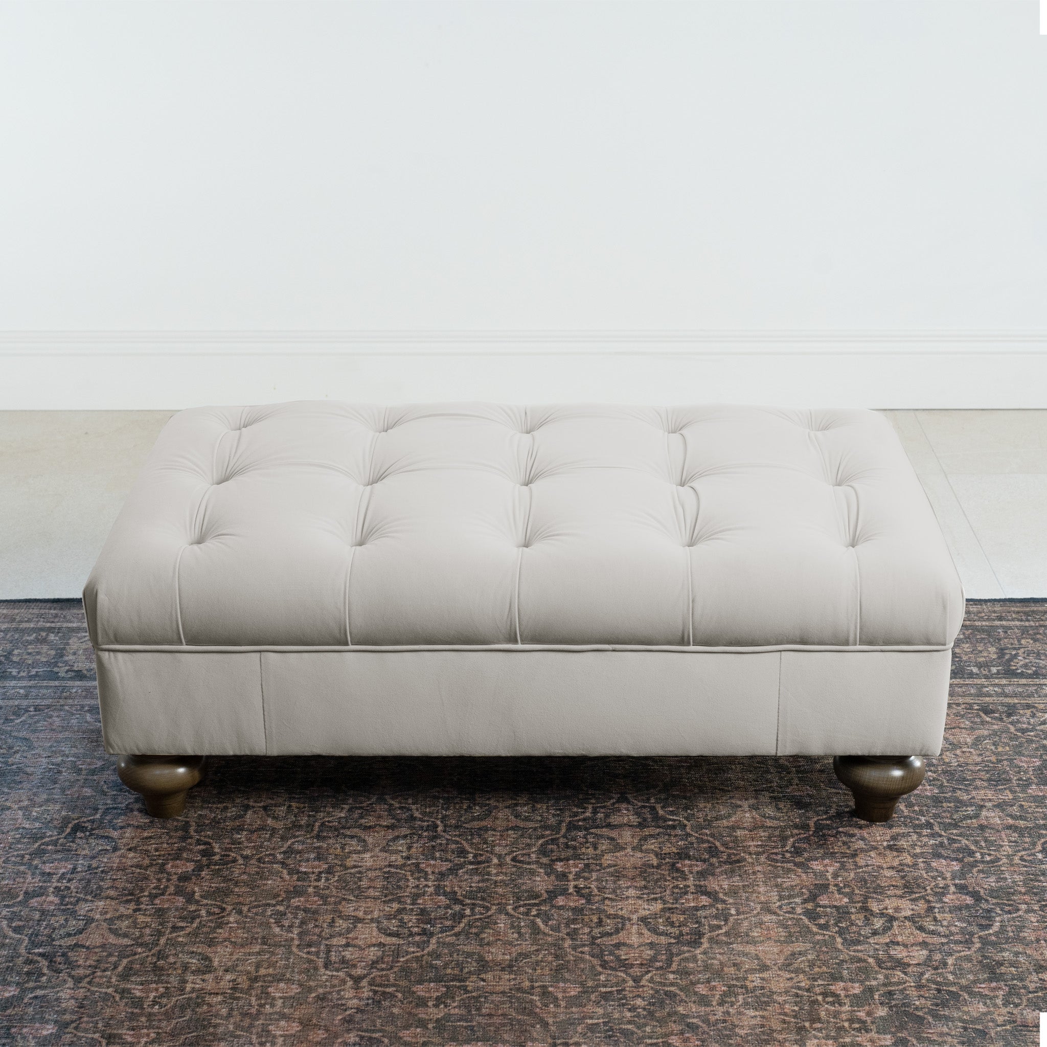 The Chesterfield Velvet Footstool in Feather Grey elegantly complements a patterned dark rug against the white wall.