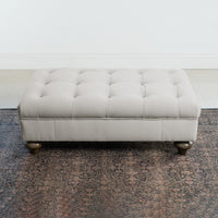 The Chesterfield Velvet Footstool in Feather Grey with wooden legs sits on a patterned rug against a white wall.