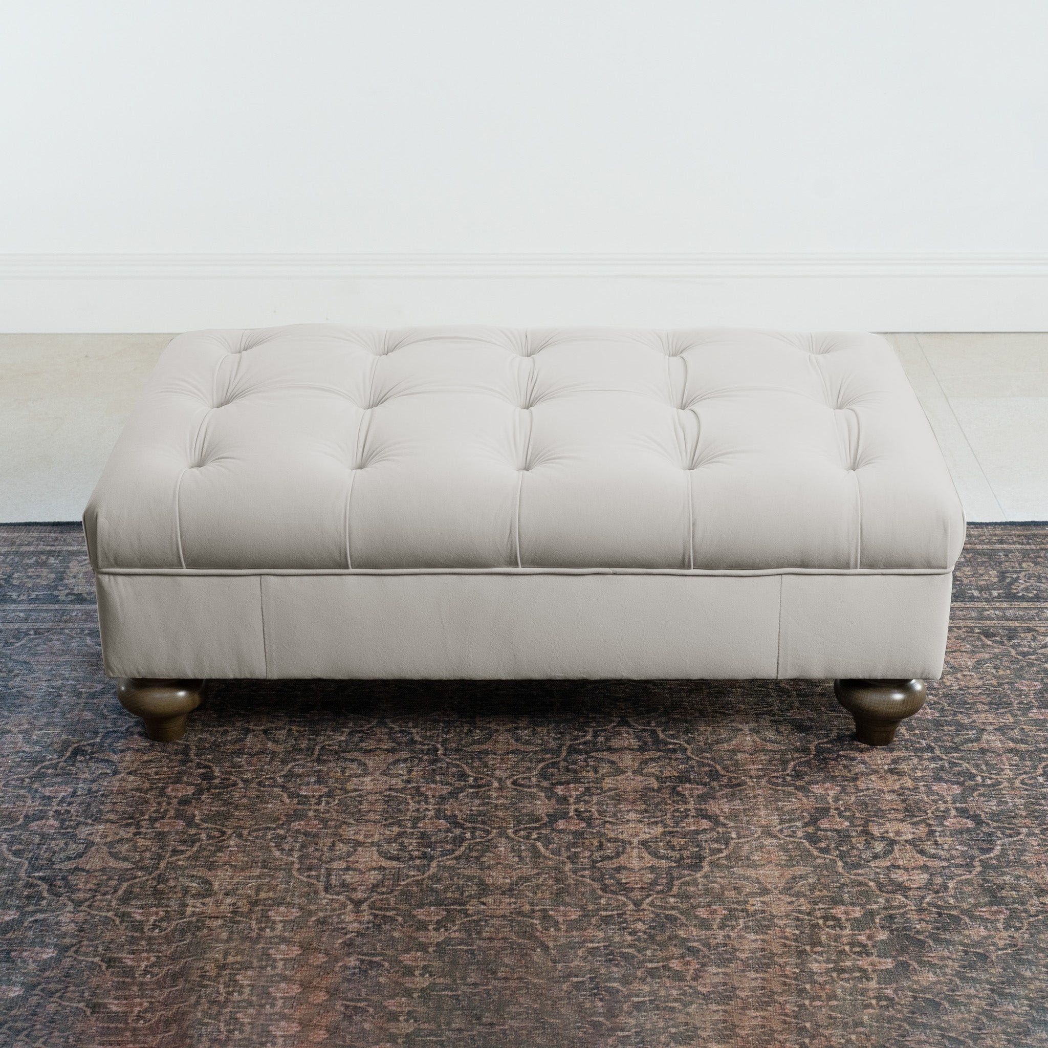 The Chesterfield Velvet Footstool in Feather Grey with wooden legs sits on a patterned rug against a white wall.