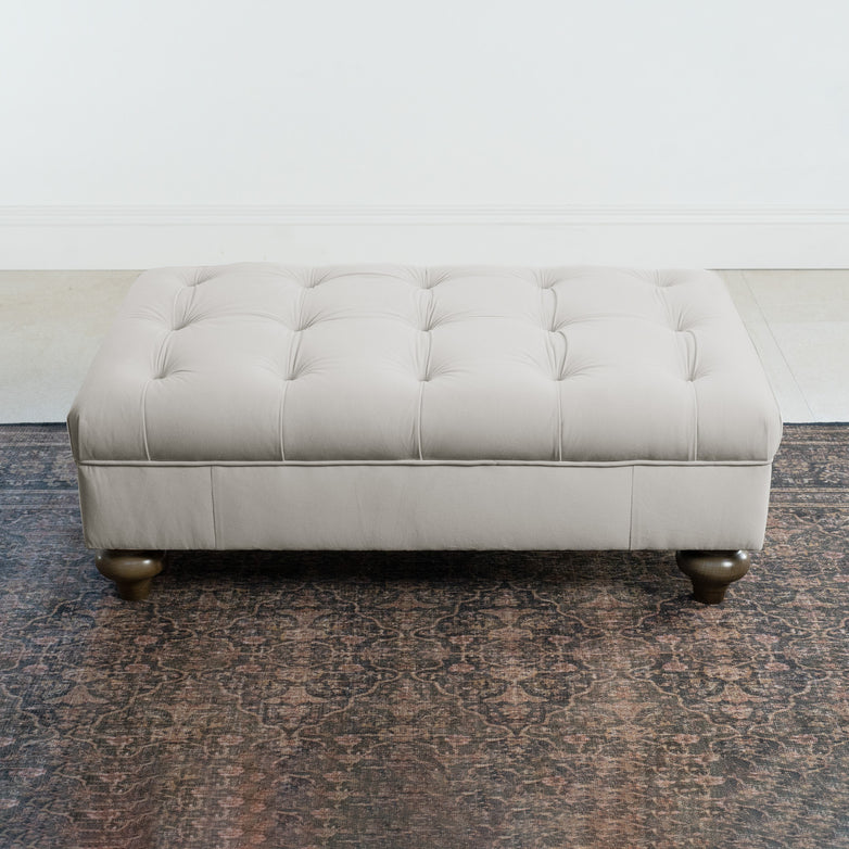 The Chesterfield Velvet Footstool in Feather Grey with wooden legs sits on a patterned rug against a white wall.