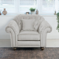 The Chesterfield Velvet Armchair in Feather Grey, meticulously handcrafted in England, pairs beautifully with the soft cushion. Positioned by a window adorned with blinds and decorative accents, it exudes an inviting sophistication.