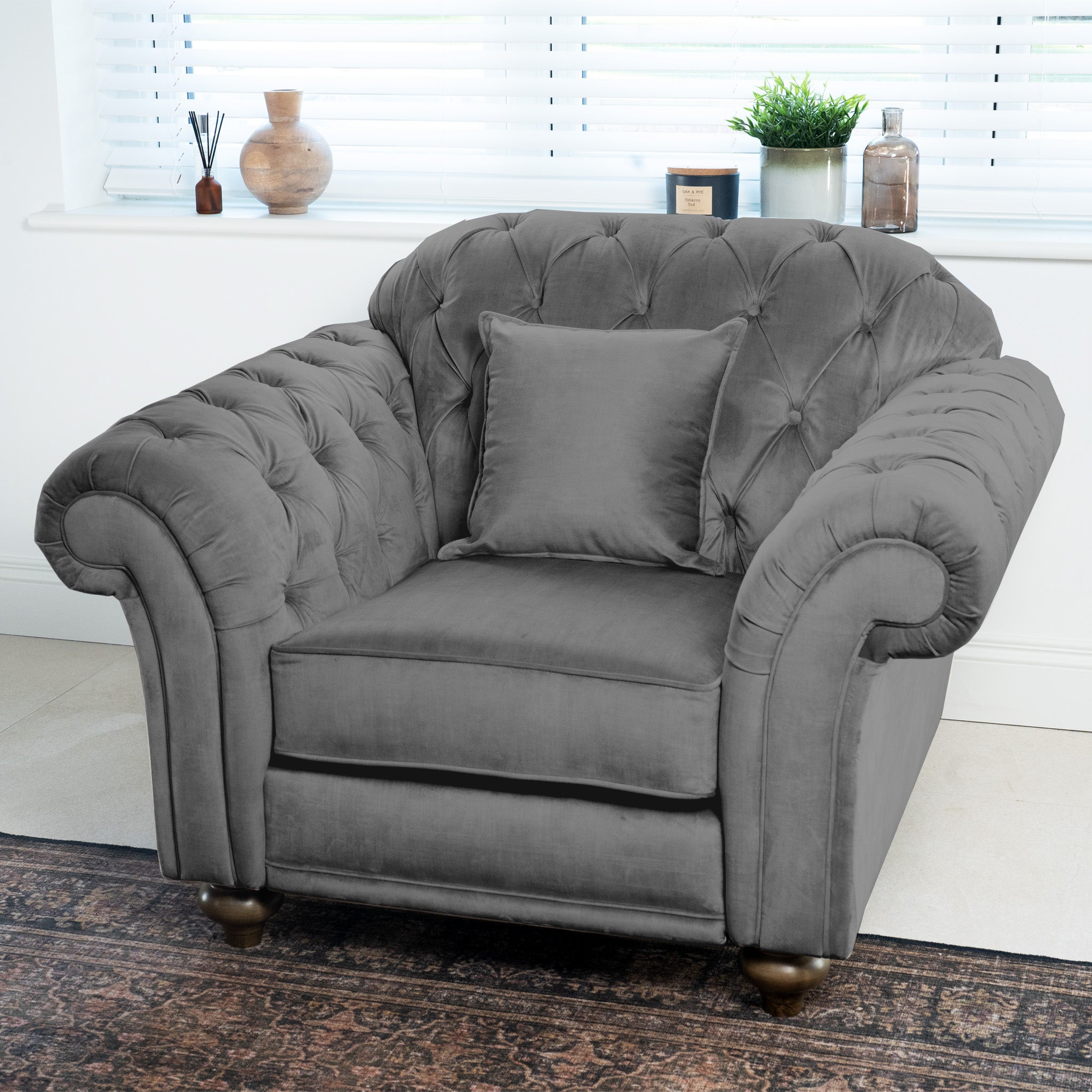 The Chesterfield Velvet Armchair in Steel Grey, featuring a matching cushion, is placed in a bright room with decorative items on the window ledge.