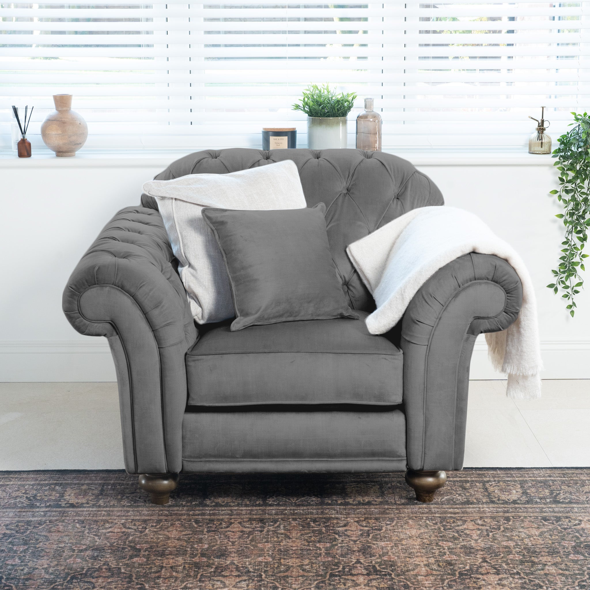 The Chesterfield Velvet Armchair in Steel Grey, paired with pillows and a blanket, is handcrafted in England. It adds elegance and warmth to any cozy room, featuring decor and a window with blinds.