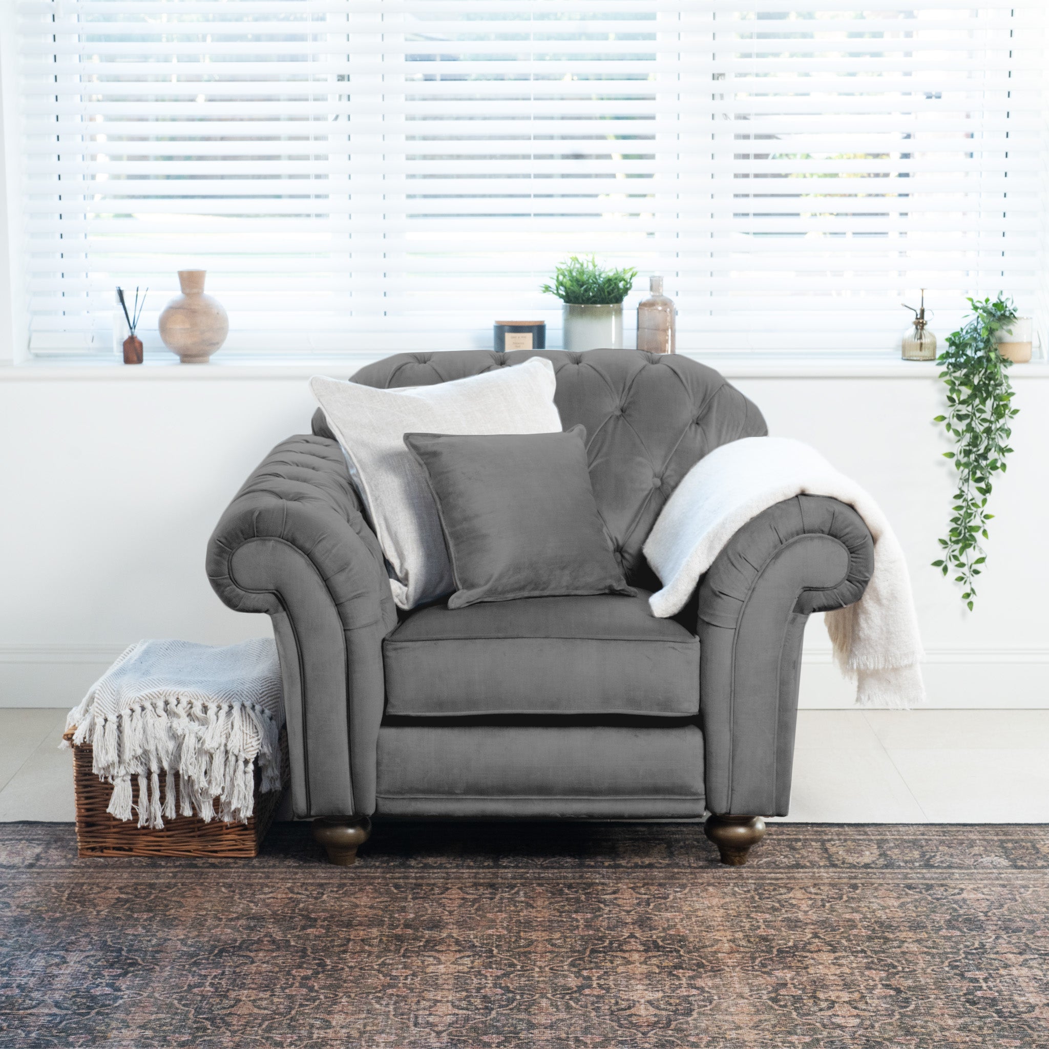 The handcrafted Chesterfield Velvet Armchair in Steel Grey, complete with pillows and a throw, sits elegantly by a window with blinds. It is made in England and surrounded by a basket and plants on the floor.