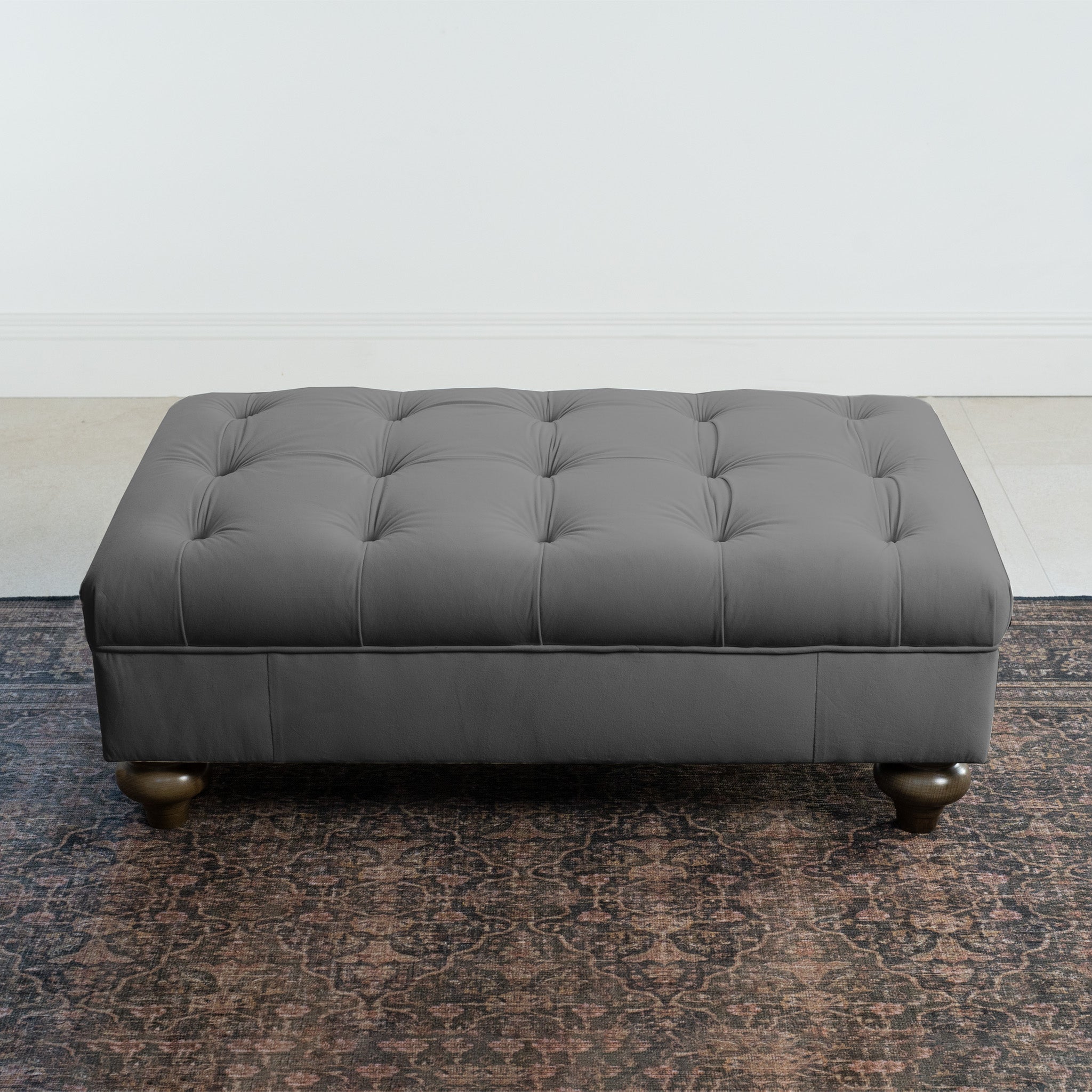 The Chesterfield Velvet Footstool in Steel Grey with tufted design rests elegantly on a patterned rug against a white wall.