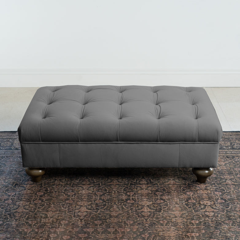 The Chesterfield Velvet Footstool in Steel Grey with tufted design rests elegantly on a patterned rug against a white wall.