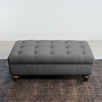 The Chesterfield Velvet Footstool in Steel Grey, handcrafted in the UK with a classic tufted design and rounded legs, looks stylish on a patterned rug against a white wall and tiled floor.