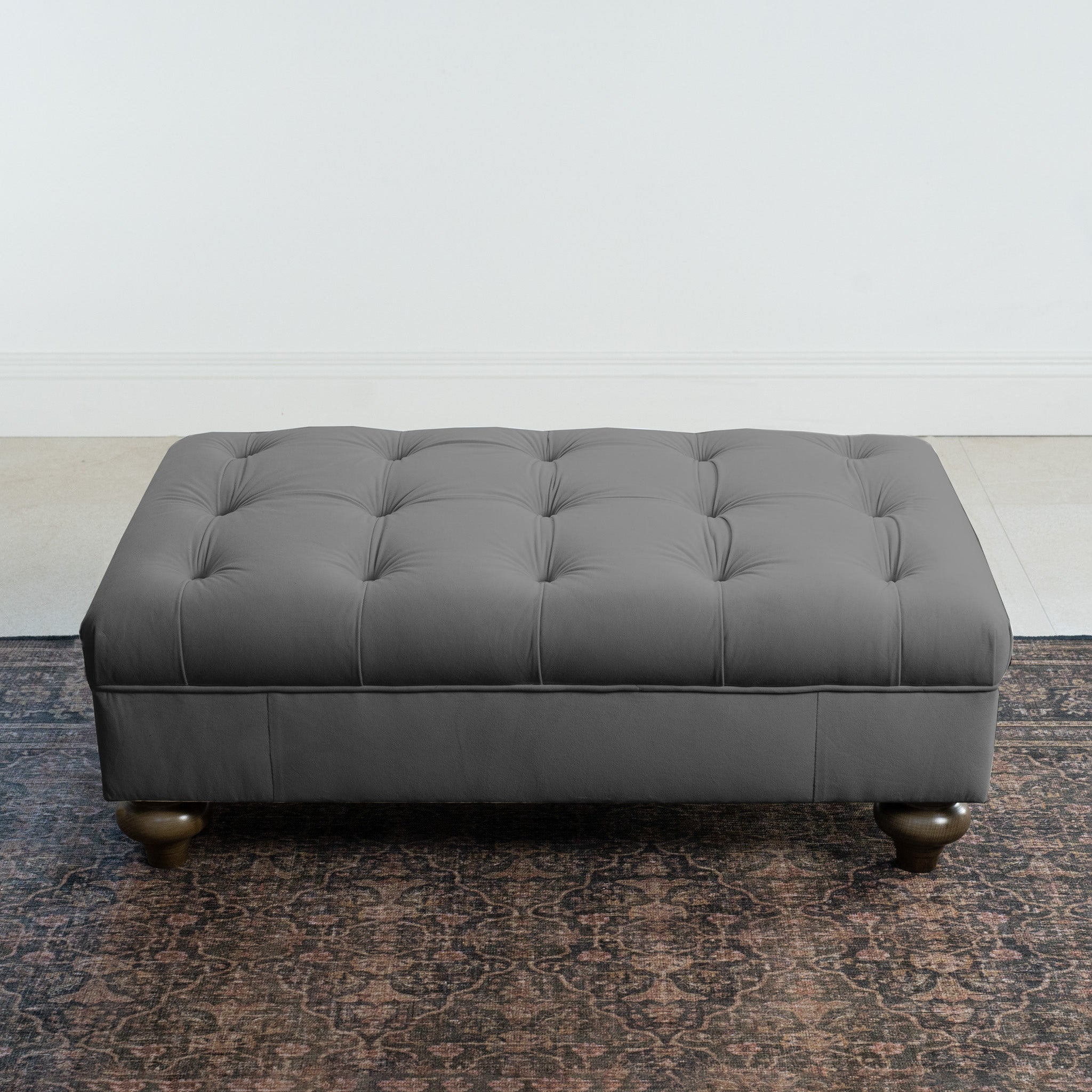 The Chesterfield Velvet Footstool in Steel Grey, handcrafted in the UK with a classic tufted design and rounded legs, looks stylish on a patterned rug against a white wall and tiled floor.