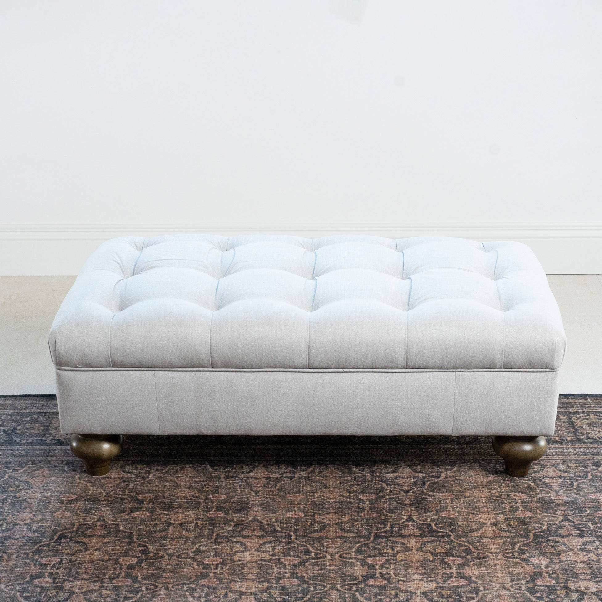 The Chesterfield Footstool in Warm Grey, featuring a tufted design and short wooden legs, sits on a patterned rug against a plain white wall.